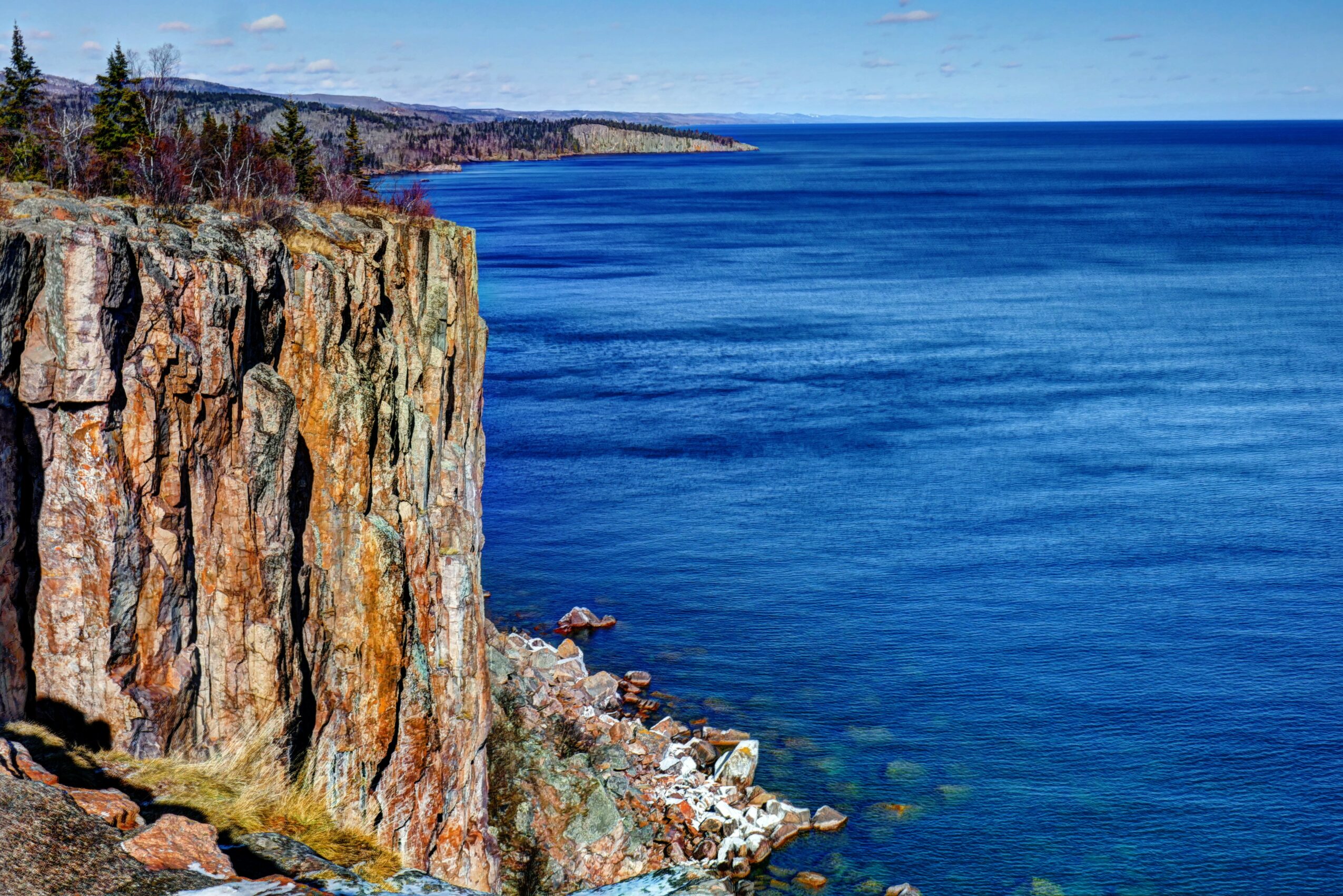 Cliff overlooking the ocean with a clear blue sky, rocky shoreline, and distant land in the background.