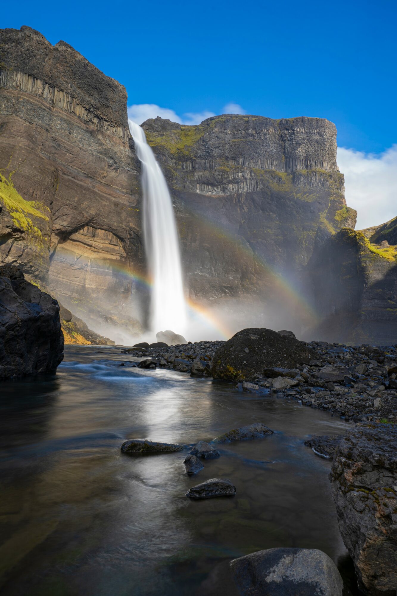 Waterfall cascading down cliffs with a rainbow in mist, river rocks in foreground, blue sky above.