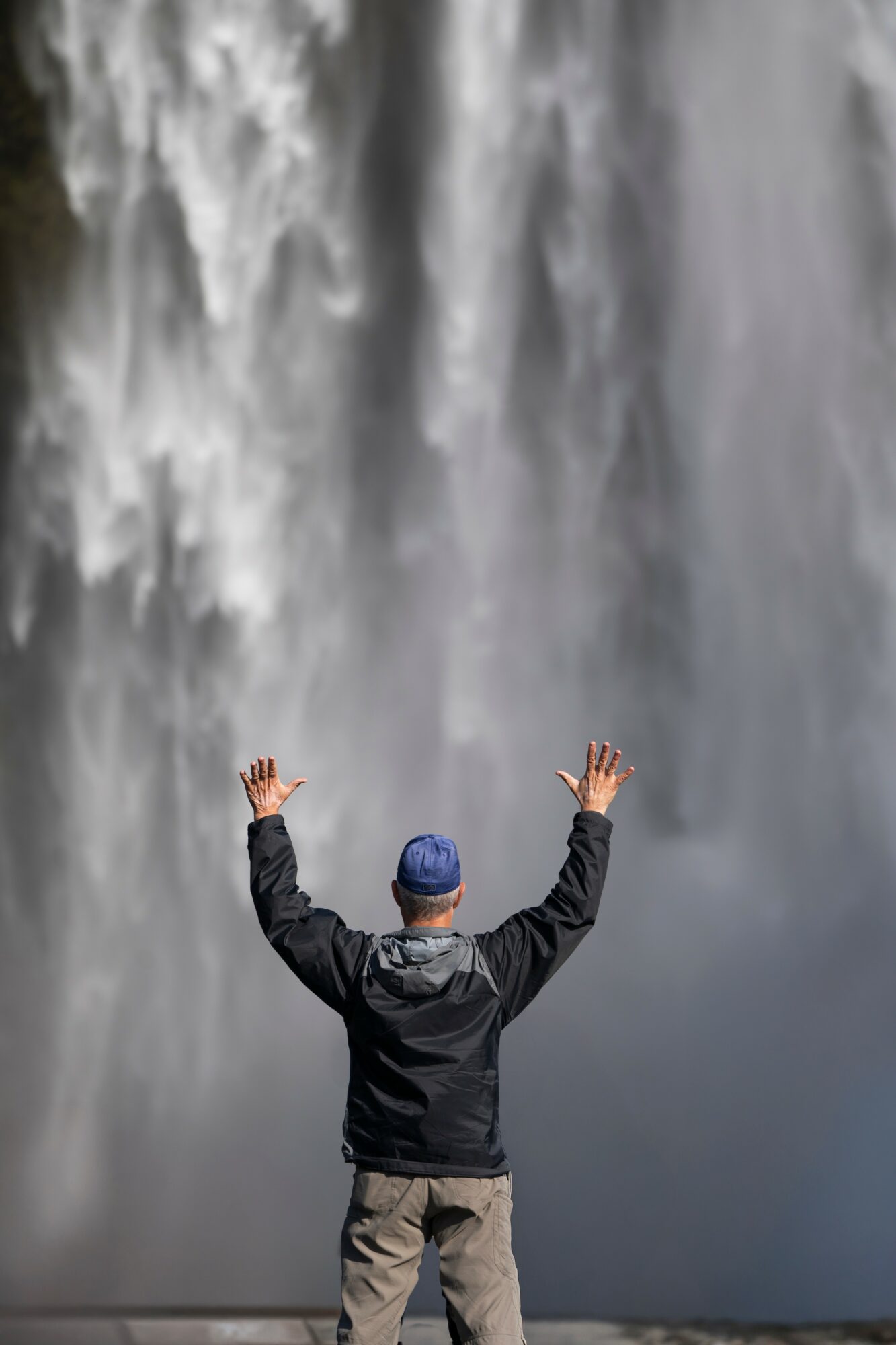 Person with arms raised facing a large waterfall, wearing a jacket and a hat, water cascading down behind them.