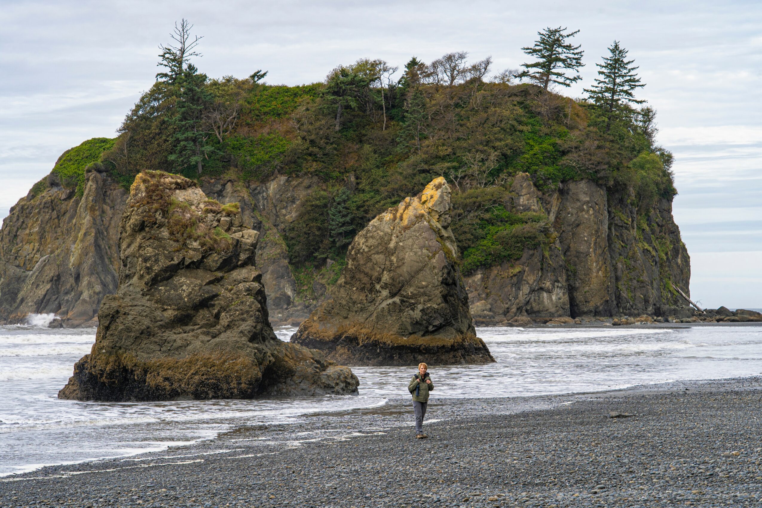 Person walking on a beach with large rock formations and a forested island in the background.