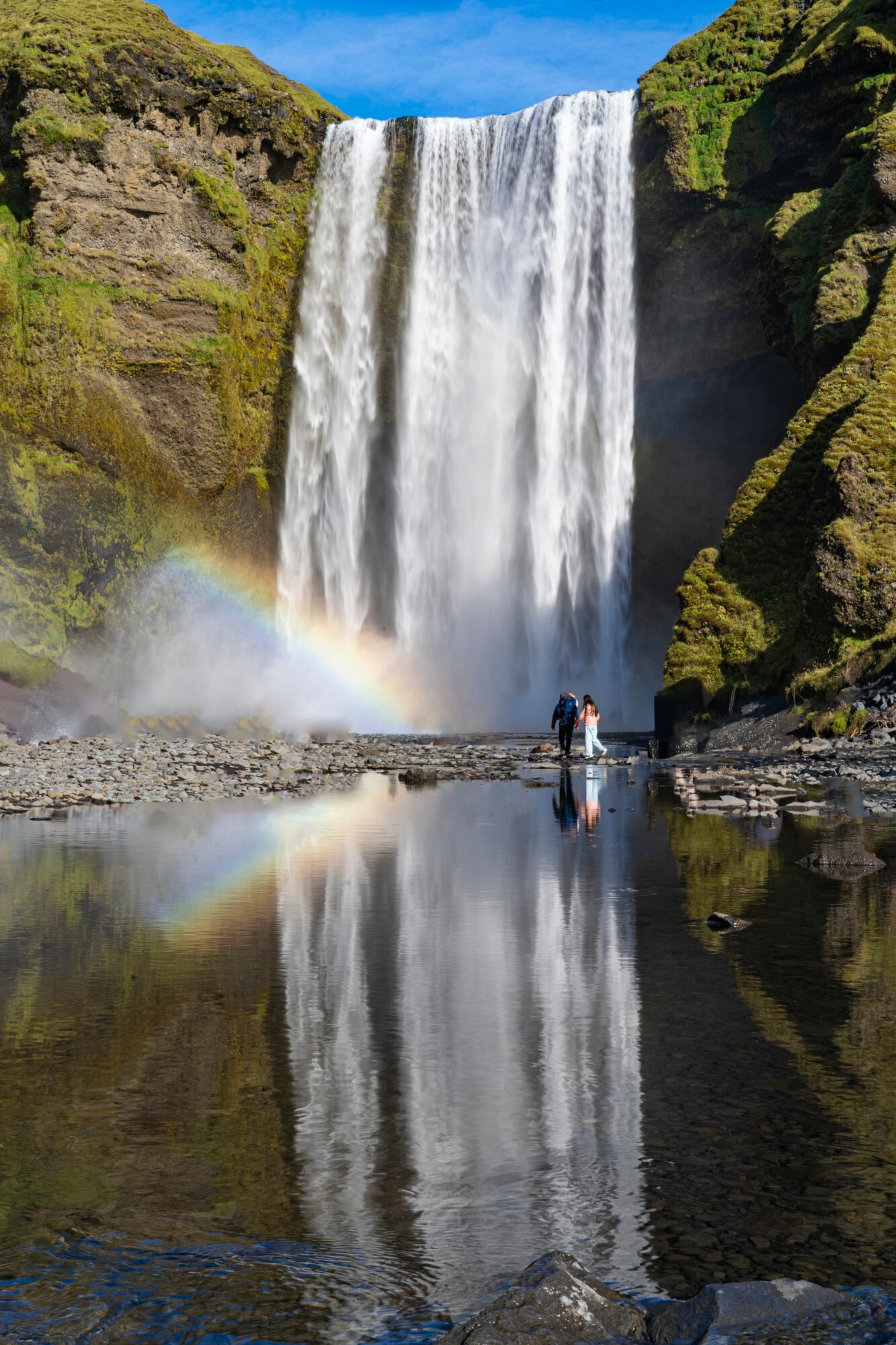 Waterfall with rainbow, person standing nearby, reflected in calm water, green cliffs, blue sky, and clouds, scenic landscape.