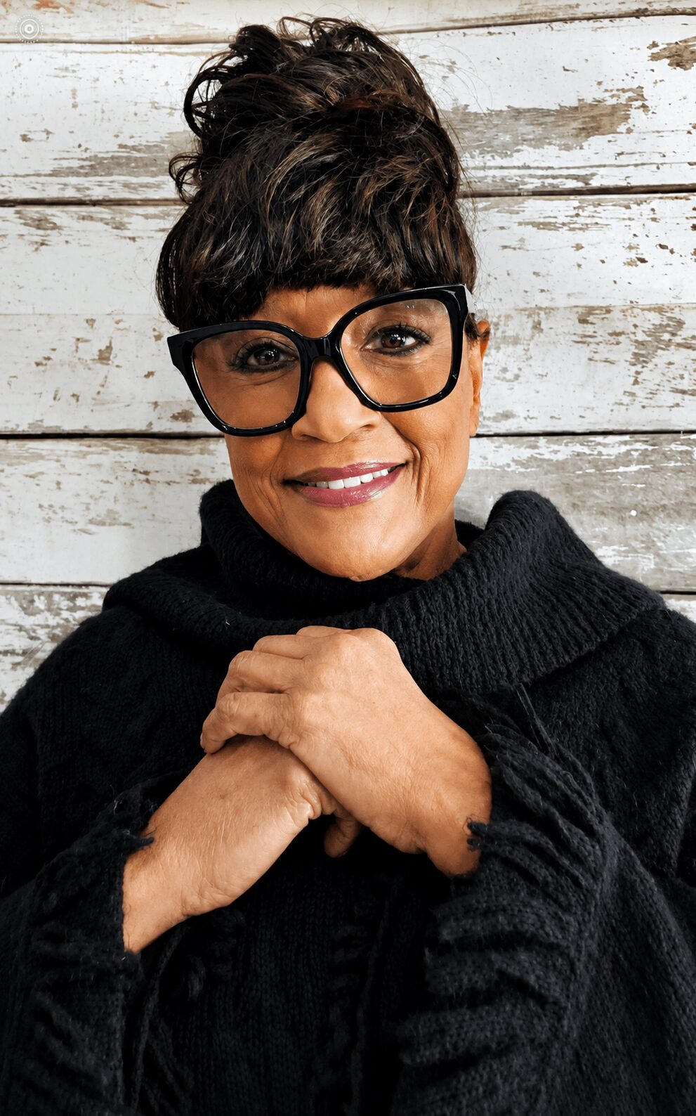 Woman with short dark hair, glasses, smiling, wearing a black sweater, against a white wooden background.