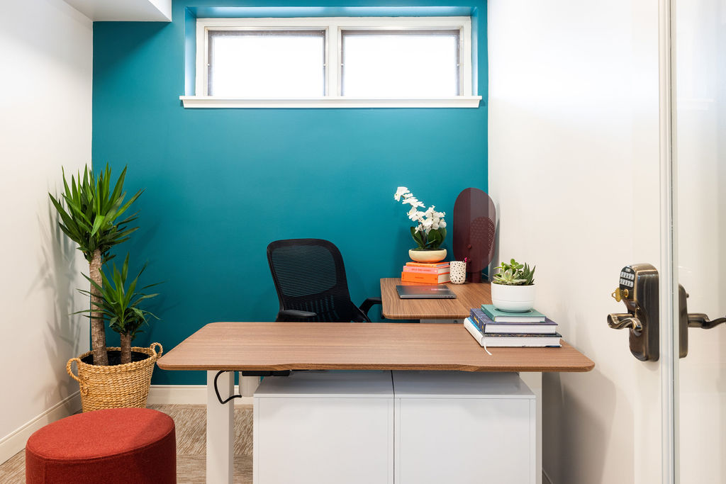 Office with a wooden desk, black chair, potted plant, and decorative items against a teal wall, window above.