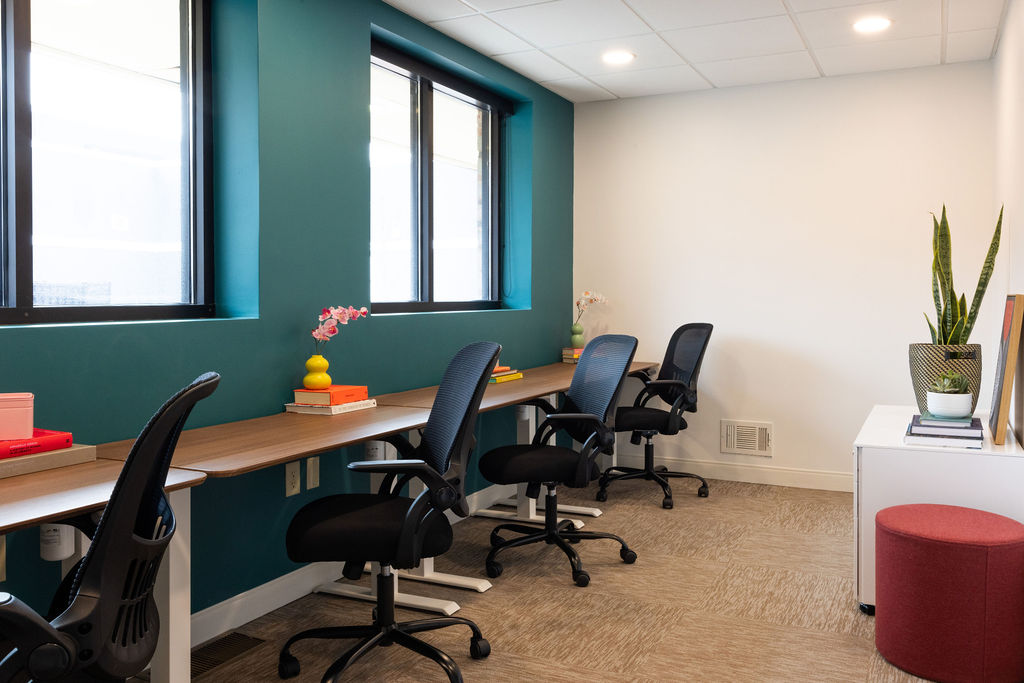 Office with a long desk, four chairs, windows, a potted plant, and a red ottoman, with beige carpet and white walls.