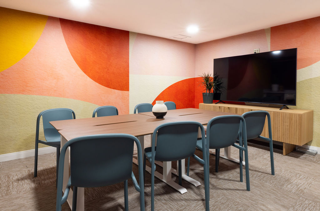 Meeting room with a wooden table, six chairs, a large TV, and colorful wall art, with a potted plant and a bowl on the table.
