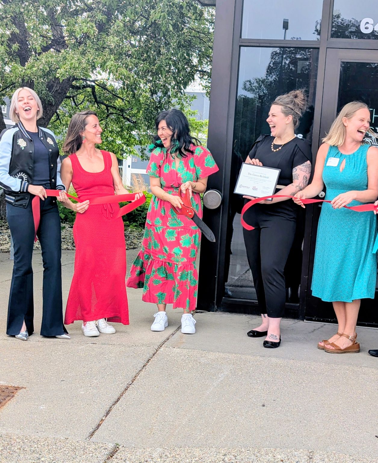 Five women standing outside, holding certificates and flowers, smiling and talking, in front of a building with glass doors.