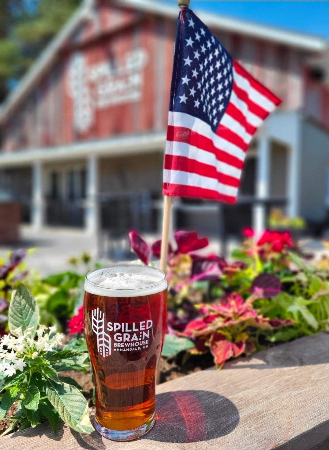 American flag on a pole, a glass of beer with spilled grain logo, and colorful flowers in front of a barn.
