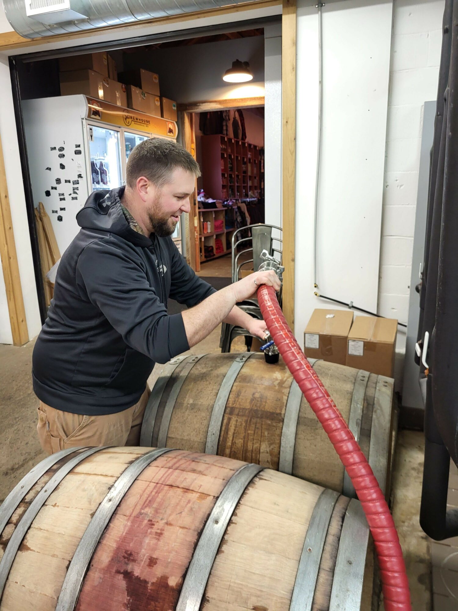 Person standing next to large wooden barrels, holding a red hose, in a room with equipment and boxes.