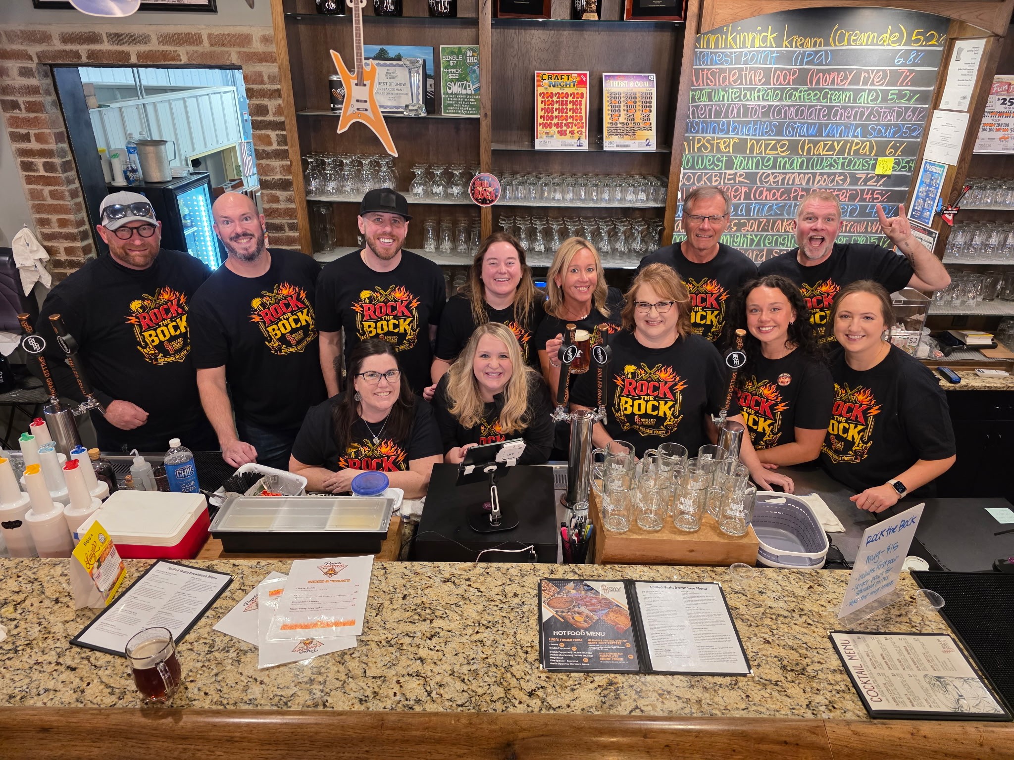 Group of people behind a bar counter, some wearing matching shirts, with glasses and a menu in front.