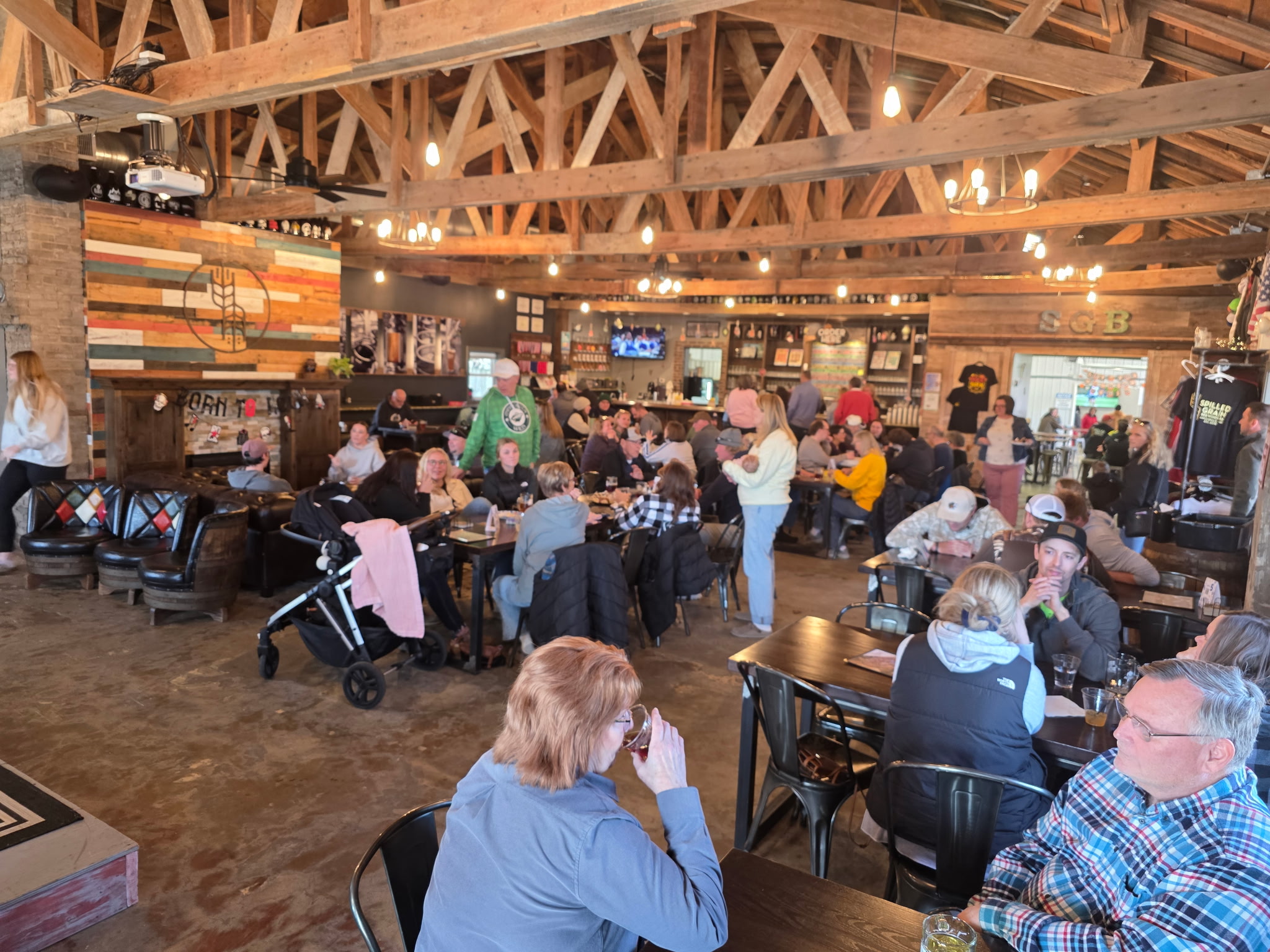 People gathered in a rustic indoor space with wooden beams, sitting at tables, socializing, and enjoying food and drinks.