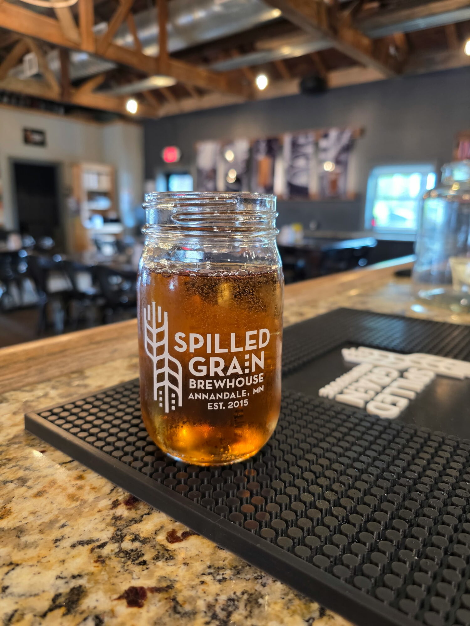 A glass jar filled with amber-colored beer on a bar counter, with a blurred bar interior in the background.