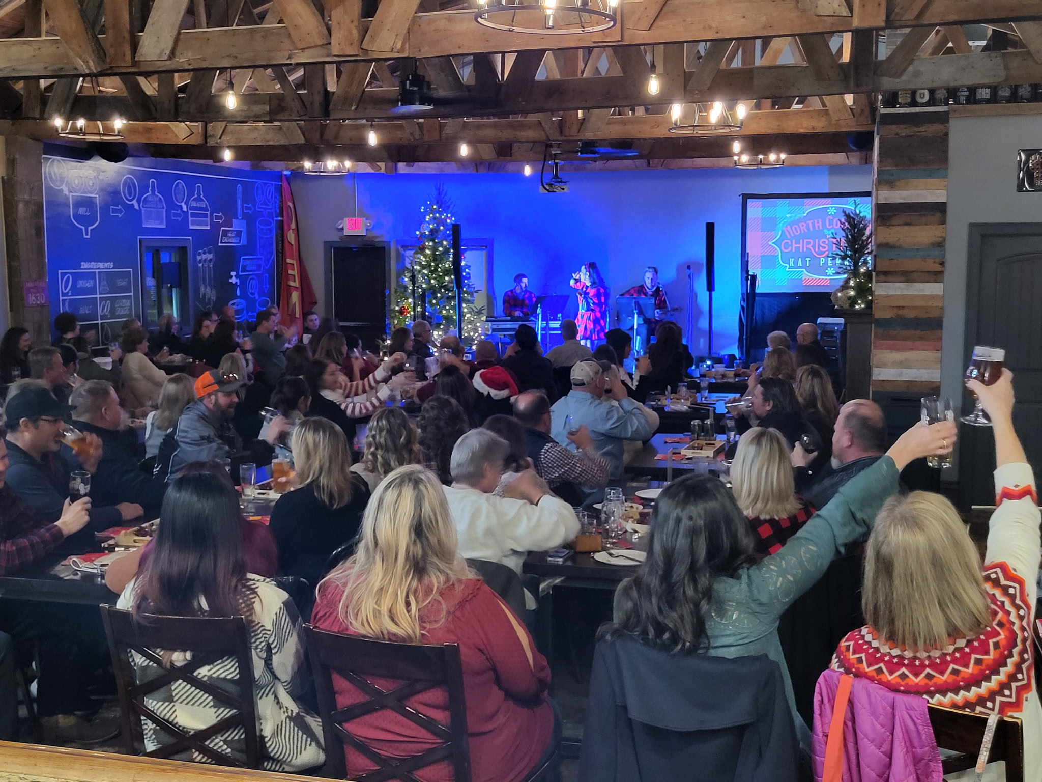 Crowd seated at tables watching a stage with performers and Christmas decorations, in a large indoor venue.