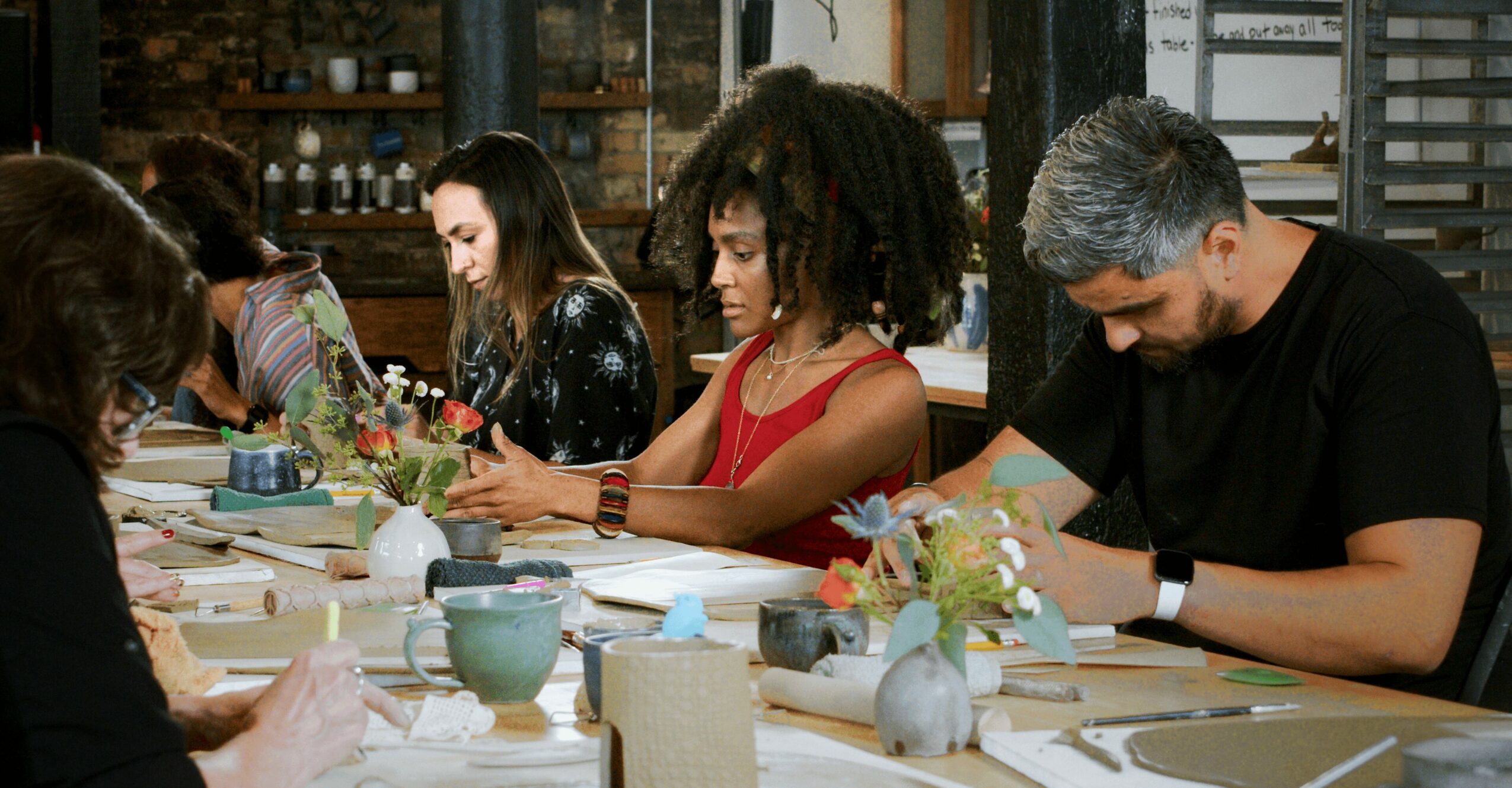 People sitting at a table with flowers and tableware, engaged in an activity, in a room with shelves and large windows.