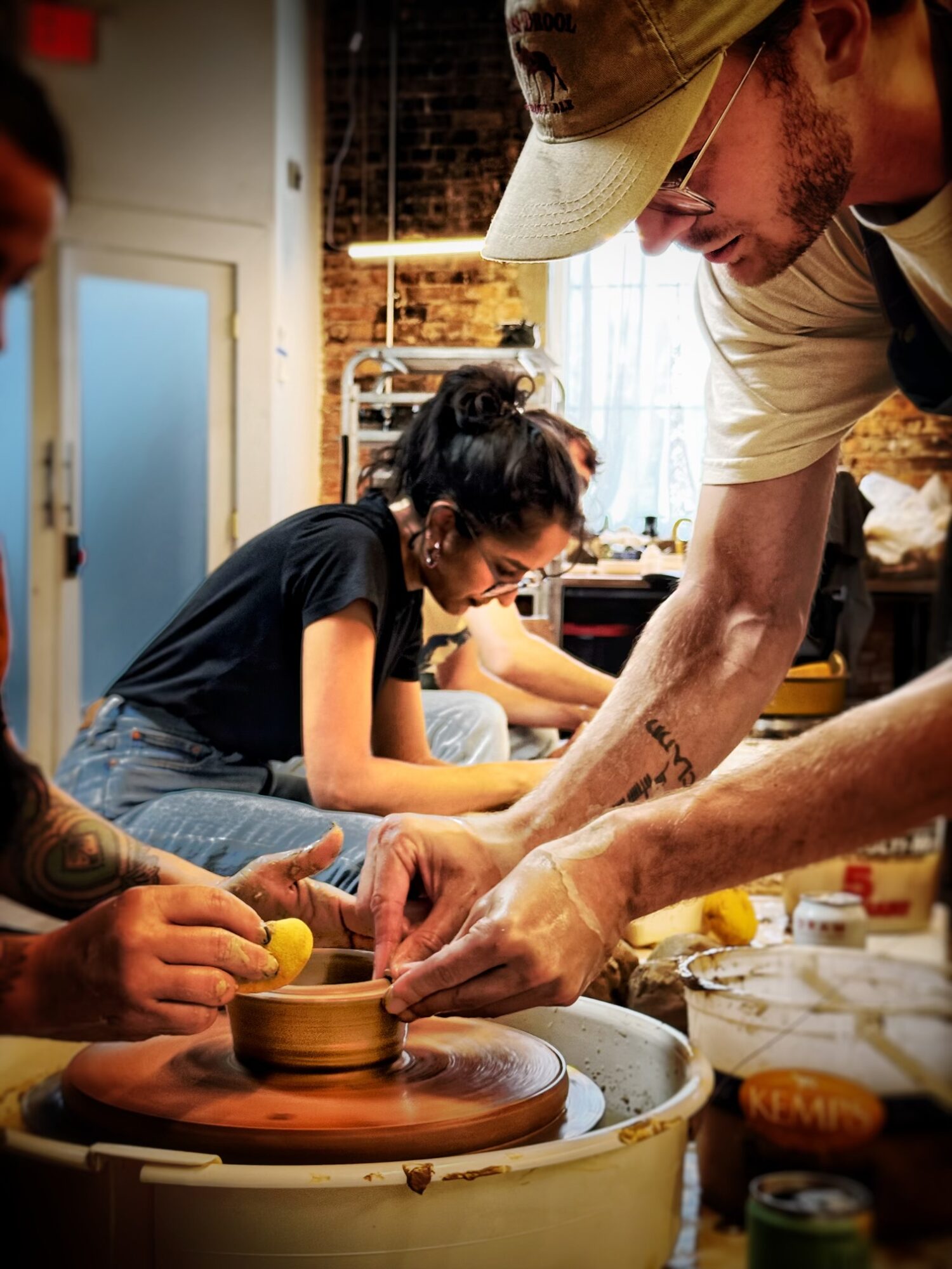 People working on pottery in a workshop, with a man guiding a woman at a pottery wheel, in a well-lit room.