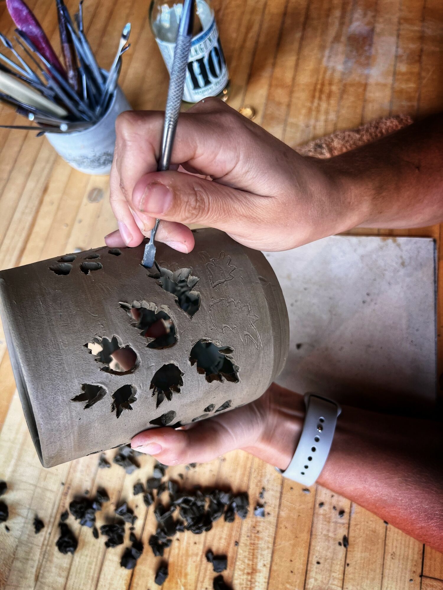 Person carving a leaf pattern into a cylindrical wooden object, with carving tools and shavings on a wooden surface.