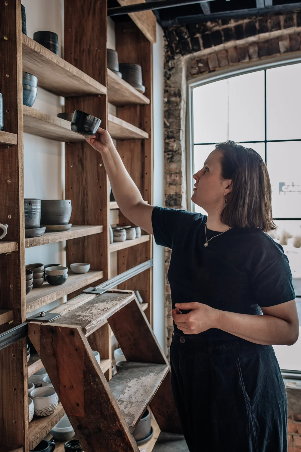 Woman reaching for a bowl on a wooden shelf in a rustic store, with a large window behind her.