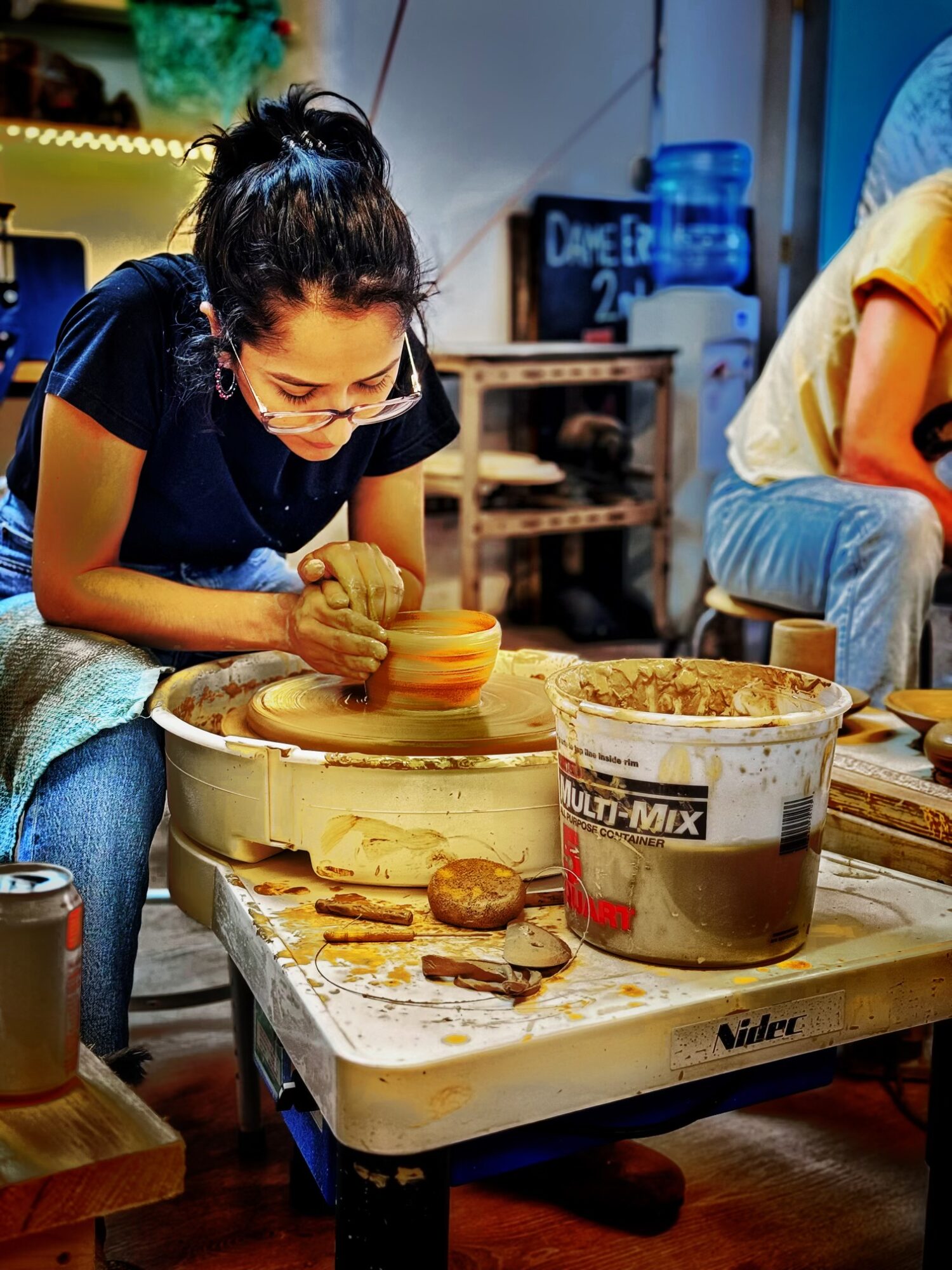 Person shaping clay on a pottery wheel in a workshop, with pottery tools and supplies nearby.