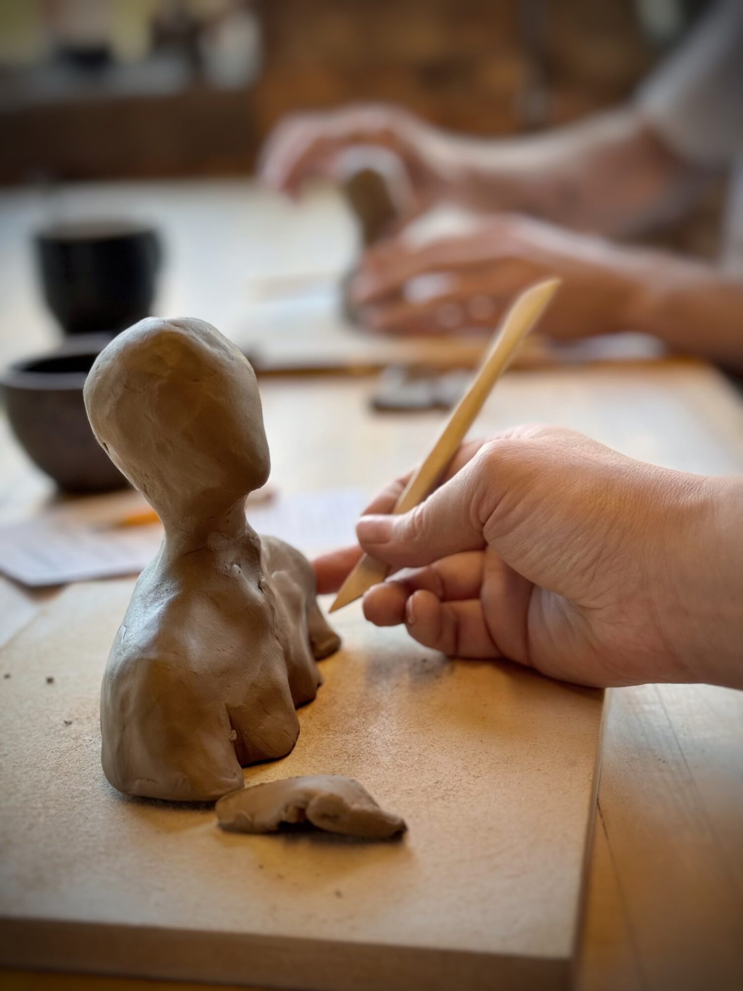 Person sculpting a clay figure with a tool, with another person in the background writing or drawing, on a wooden table.