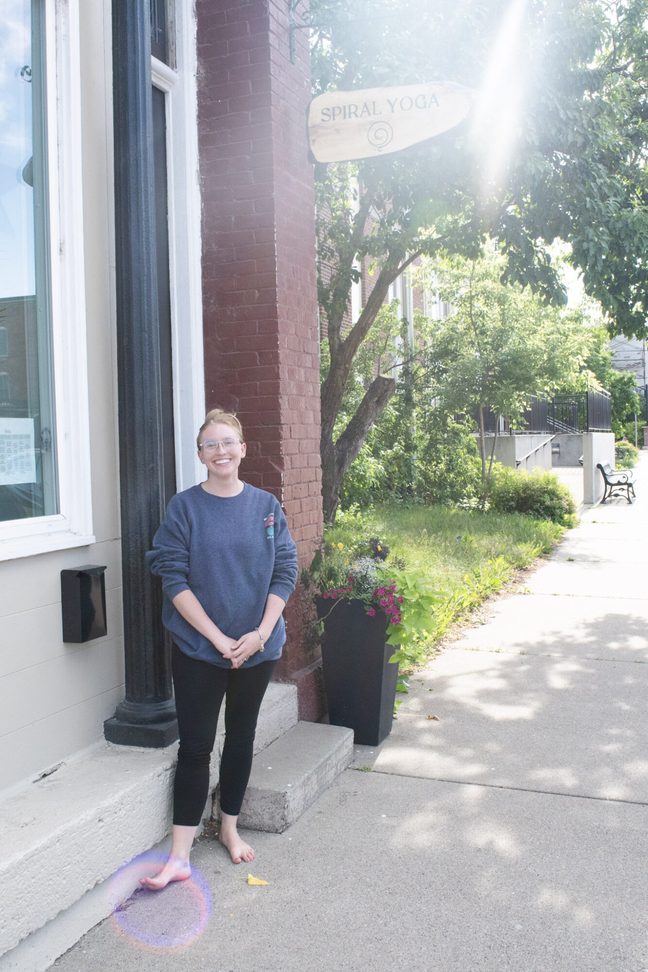 Person standing on sidewalk near building with window, holding a paper, with trees and sunlight in background.