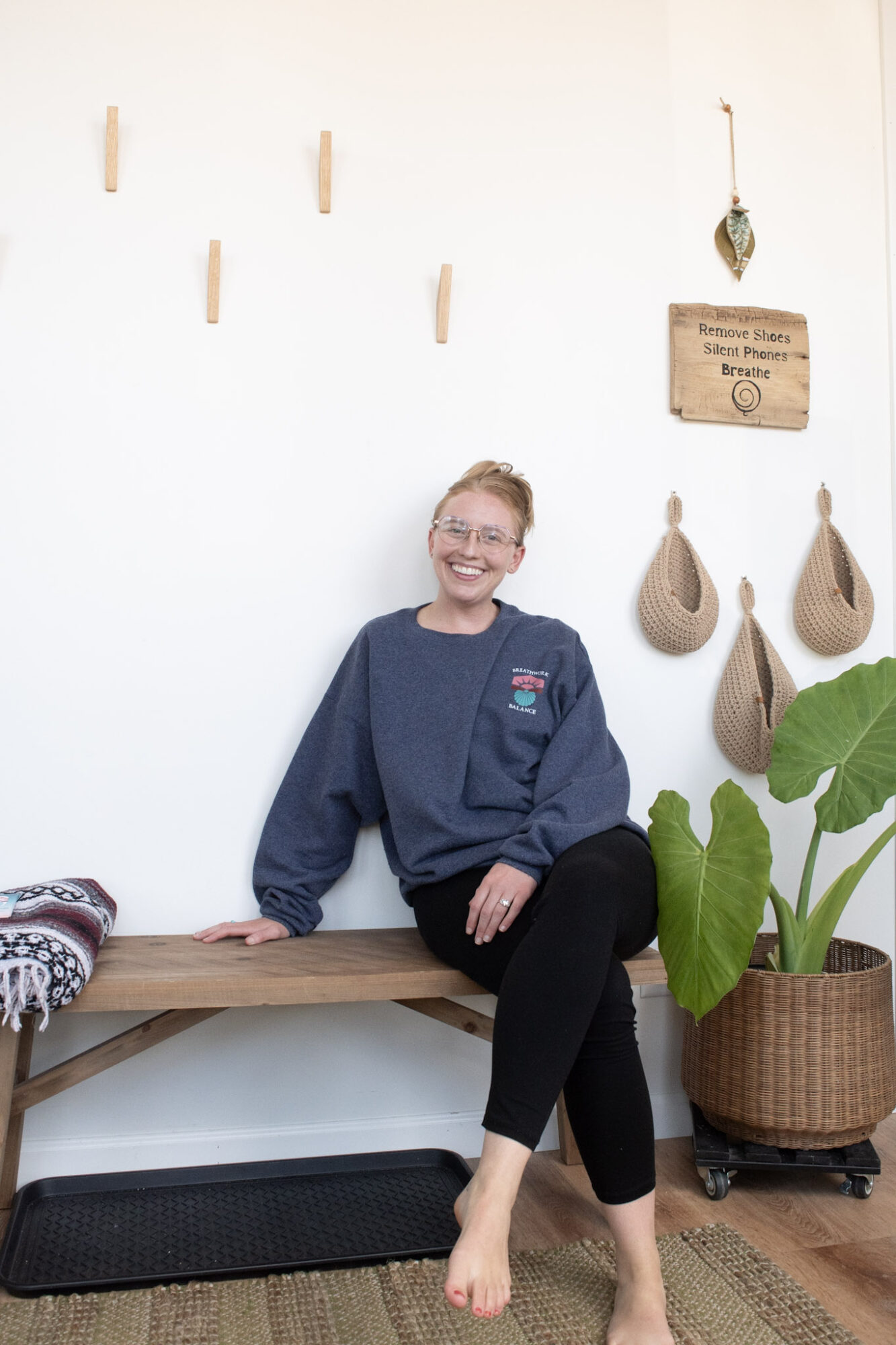 Person with glasses and a dark sweatshirt sitting on a wooden bench, smiling, with hanging decorative items and a plant nearby.