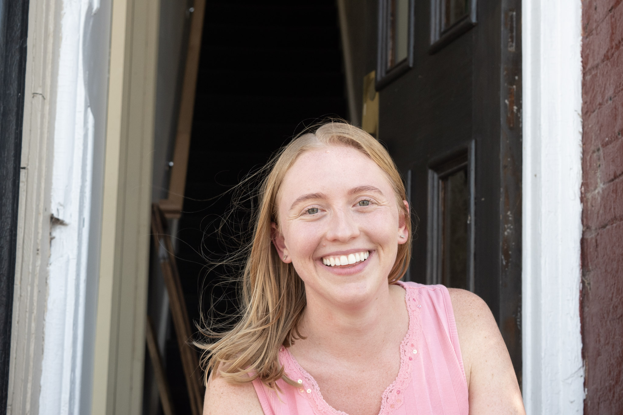 Young woman with long blonde hair smiling, wearing a pink sleeveless top, standing in front of a door.