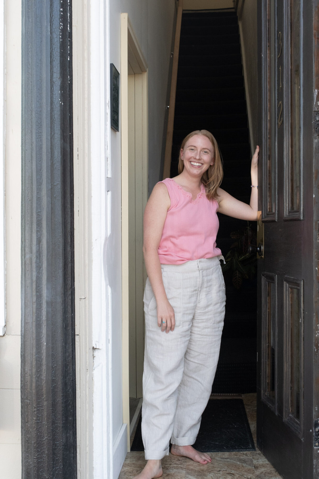 Woman standing in doorway, smiling, wearing pink top and white pants, with stairs visible in background.
