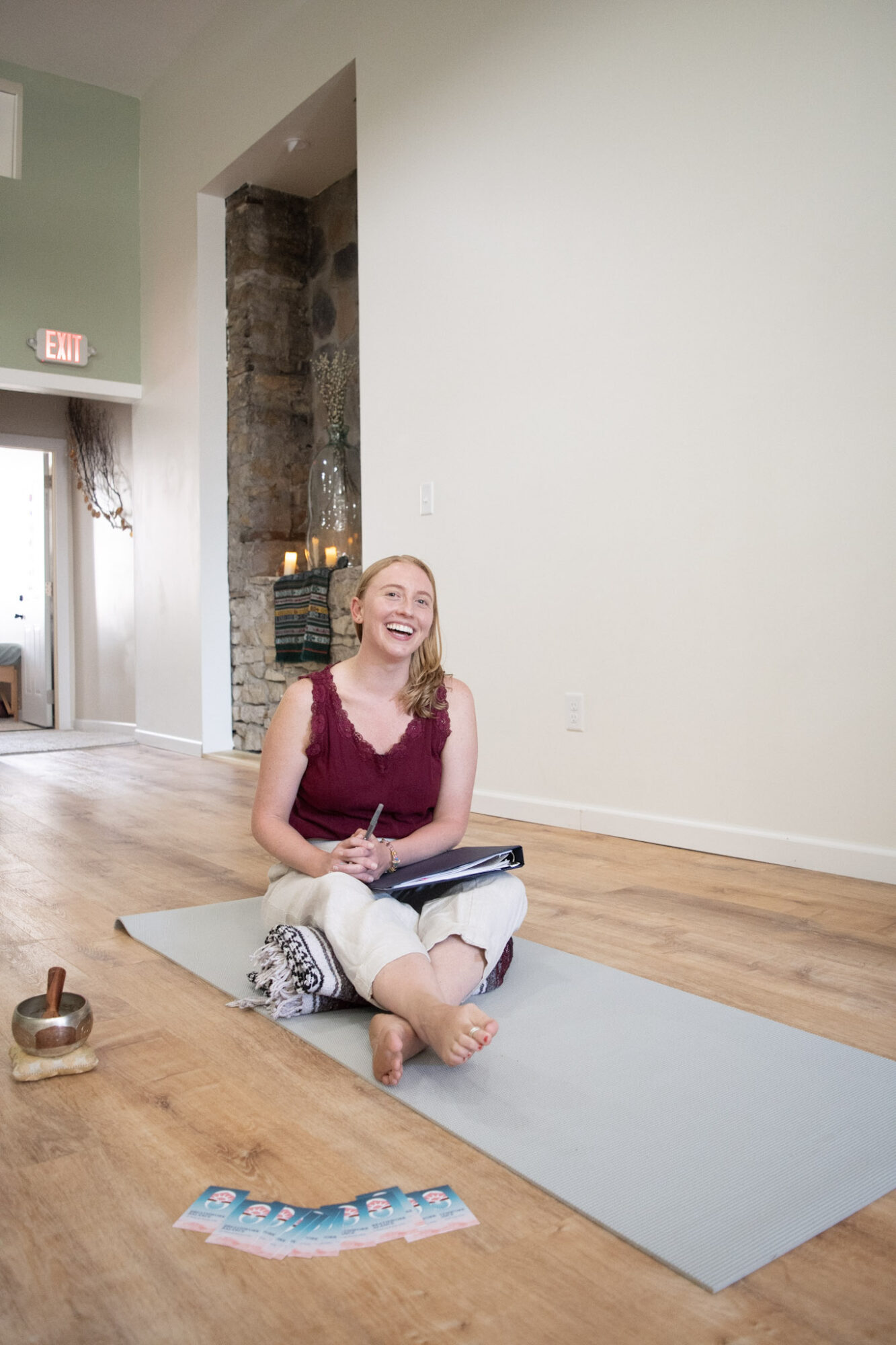 Woman sitting cross-legged on a yoga mat, smiling, in a spacious room with wooden floor and white walls.