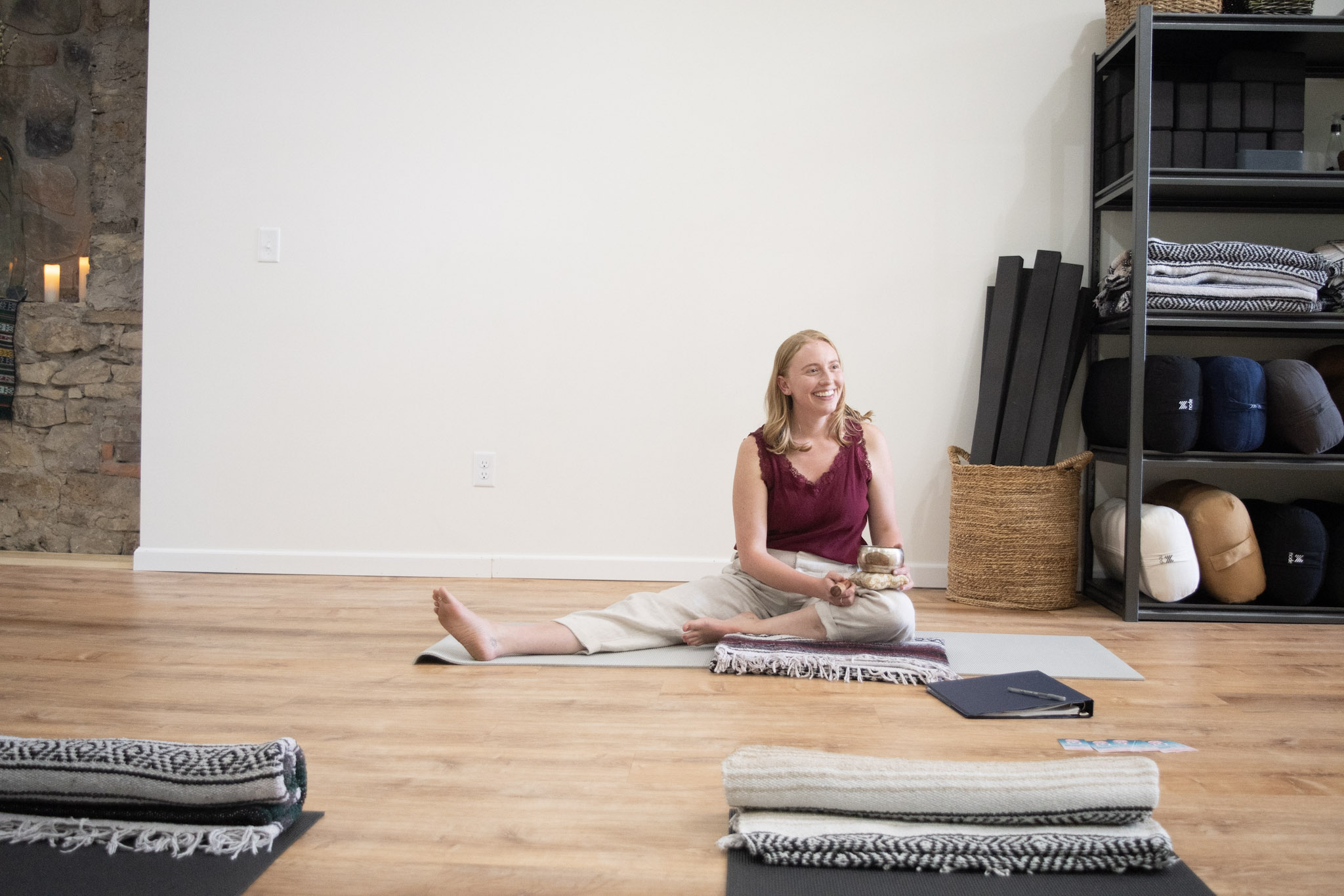 Woman sitting on the floor in a yoga pose, smiling, in a room with wooden floor and shelves, with yoga mats and books nearby.