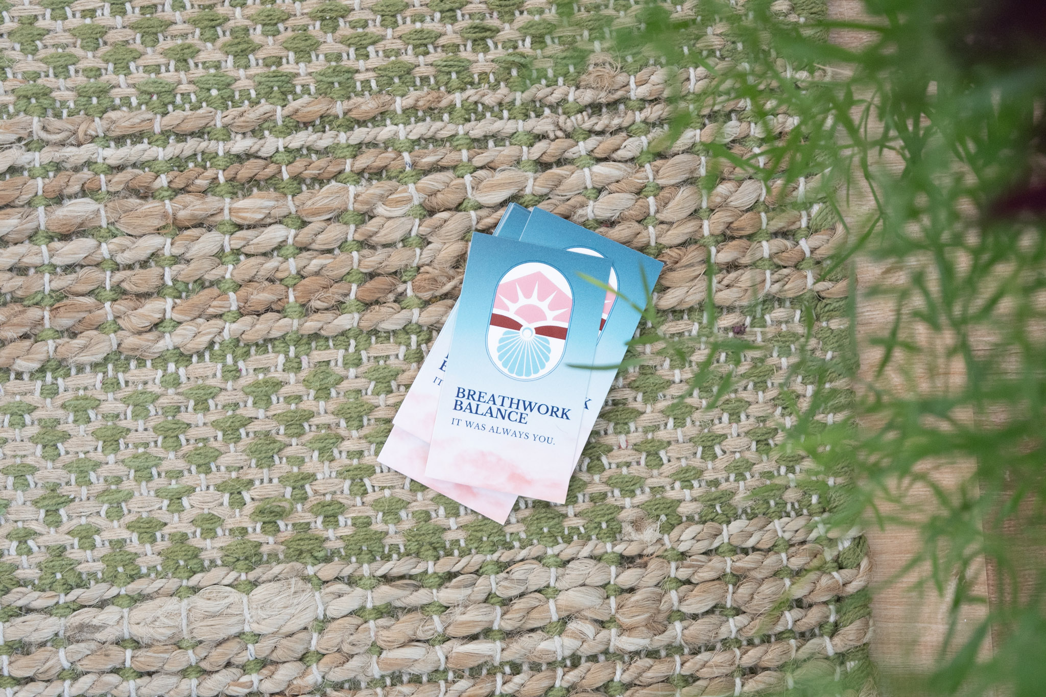 A tea bag with a blue and pink label on a woven surface, partially covered by green foliage.