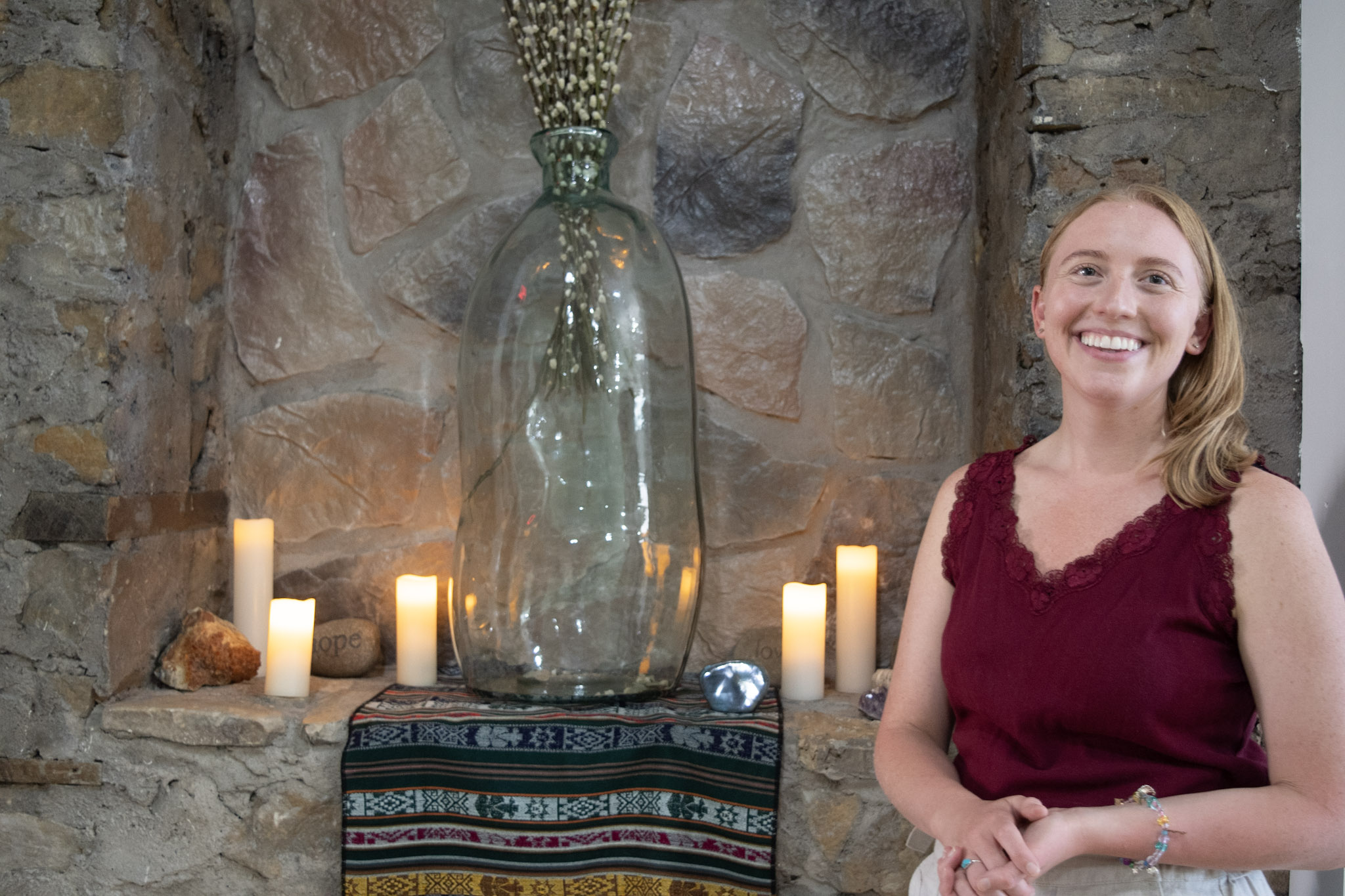 Smiling woman in a burgundy dress standing in front of a stone wall with candles and a large glass vase.