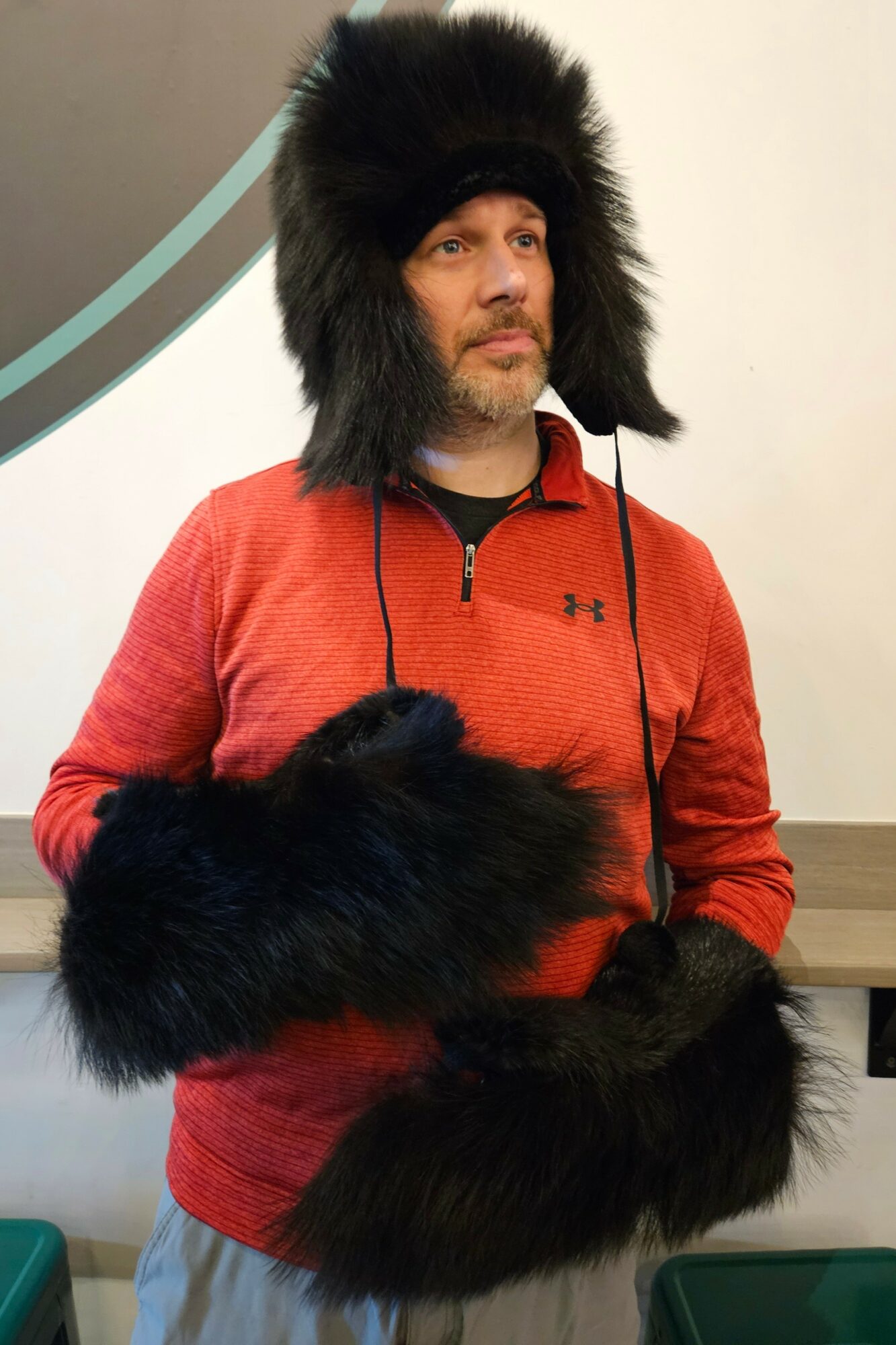 Man wearing a large black furry hat and matching mittens, red zip-up jacket, standing indoors against a white wall.