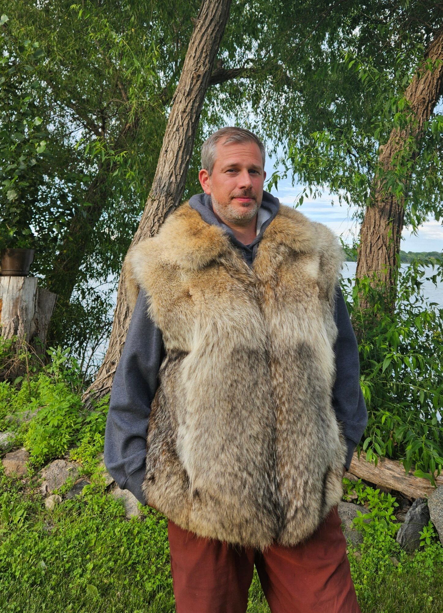 Man standing outdoors near trees, wearing a large fur vest and maroon pants, with greenery and water in background.