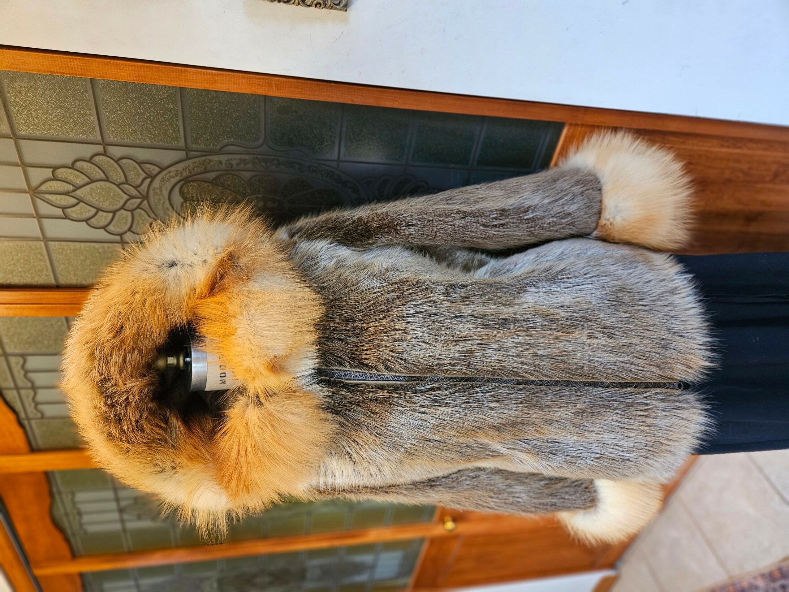 Fur coat with a large, fluffy hood, hanging on a hook against a tiled wall and wooden panel, with a person partially visible.