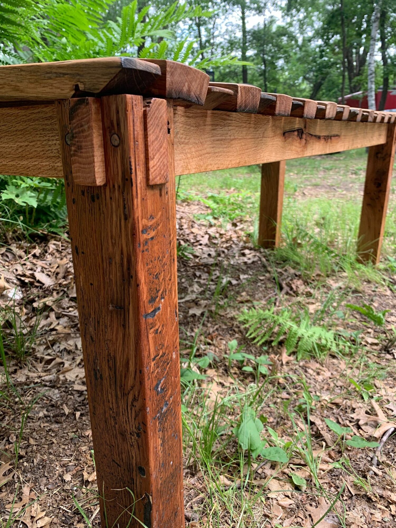 Wooden bench outdoors on grass with trees in background, viewed from the side, showing the bench's length and structure.
