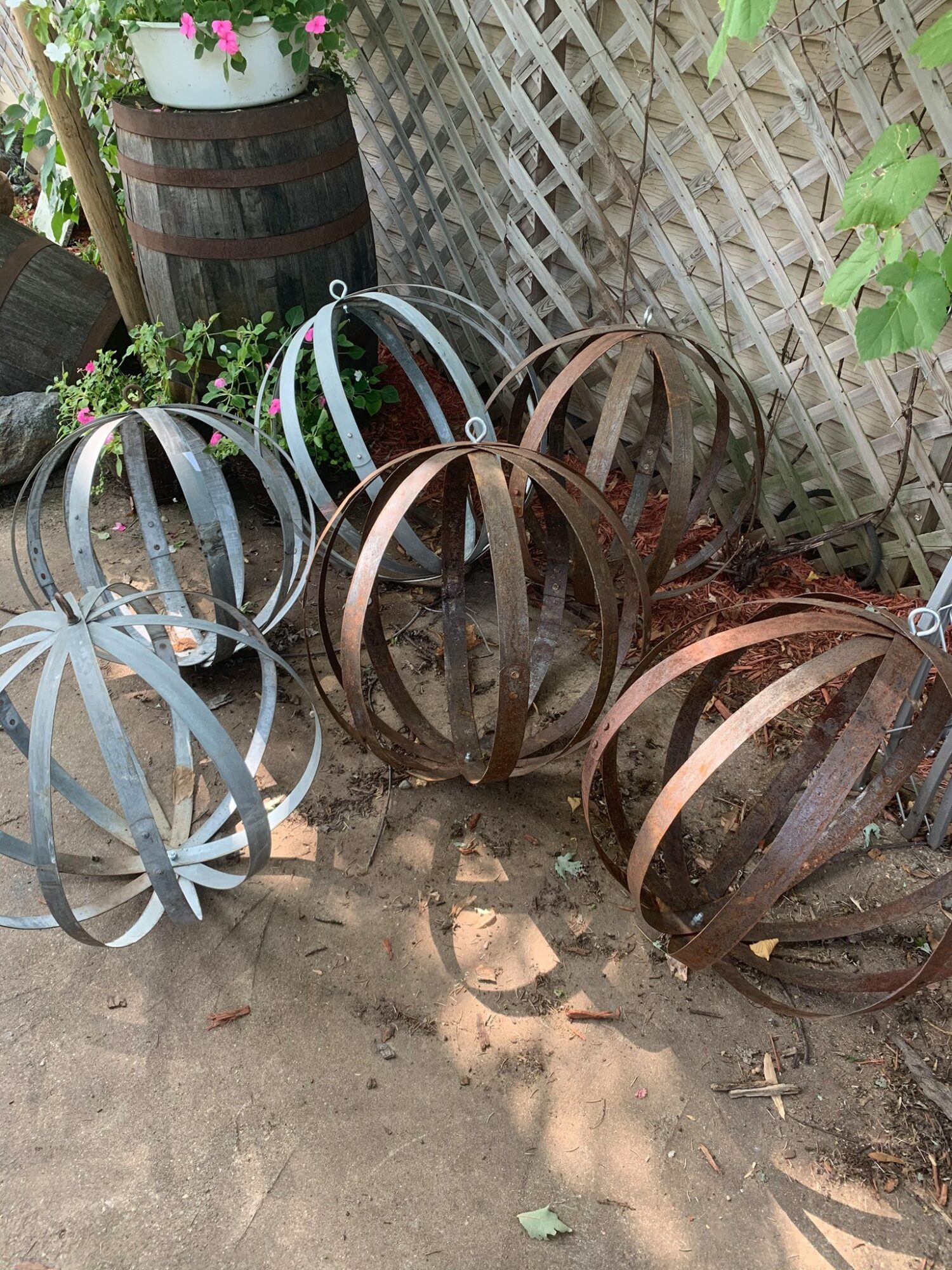 Several metal garden ornaments shaped like spheres with open slats, placed on dirt near a wooden fence and plants.