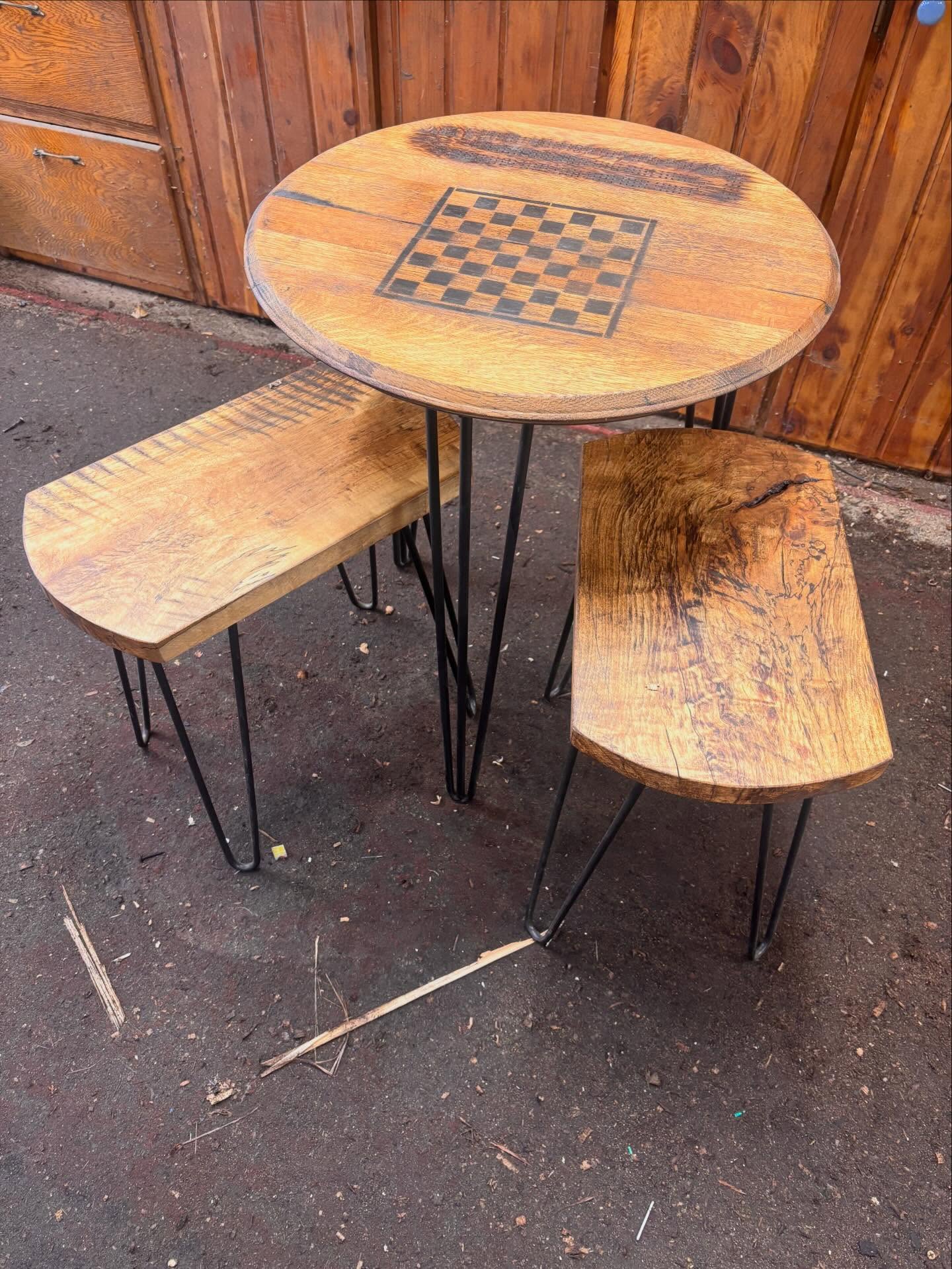 Round wooden table with a chessboard, two wooden benches with metal legs, outdoor setting with wooden fence background.