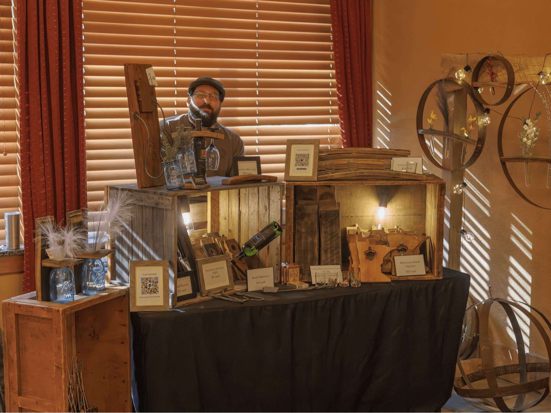 Man taking a selfie in a mirror behind a display table with framed pictures and decorative items, sunlight through blinds.