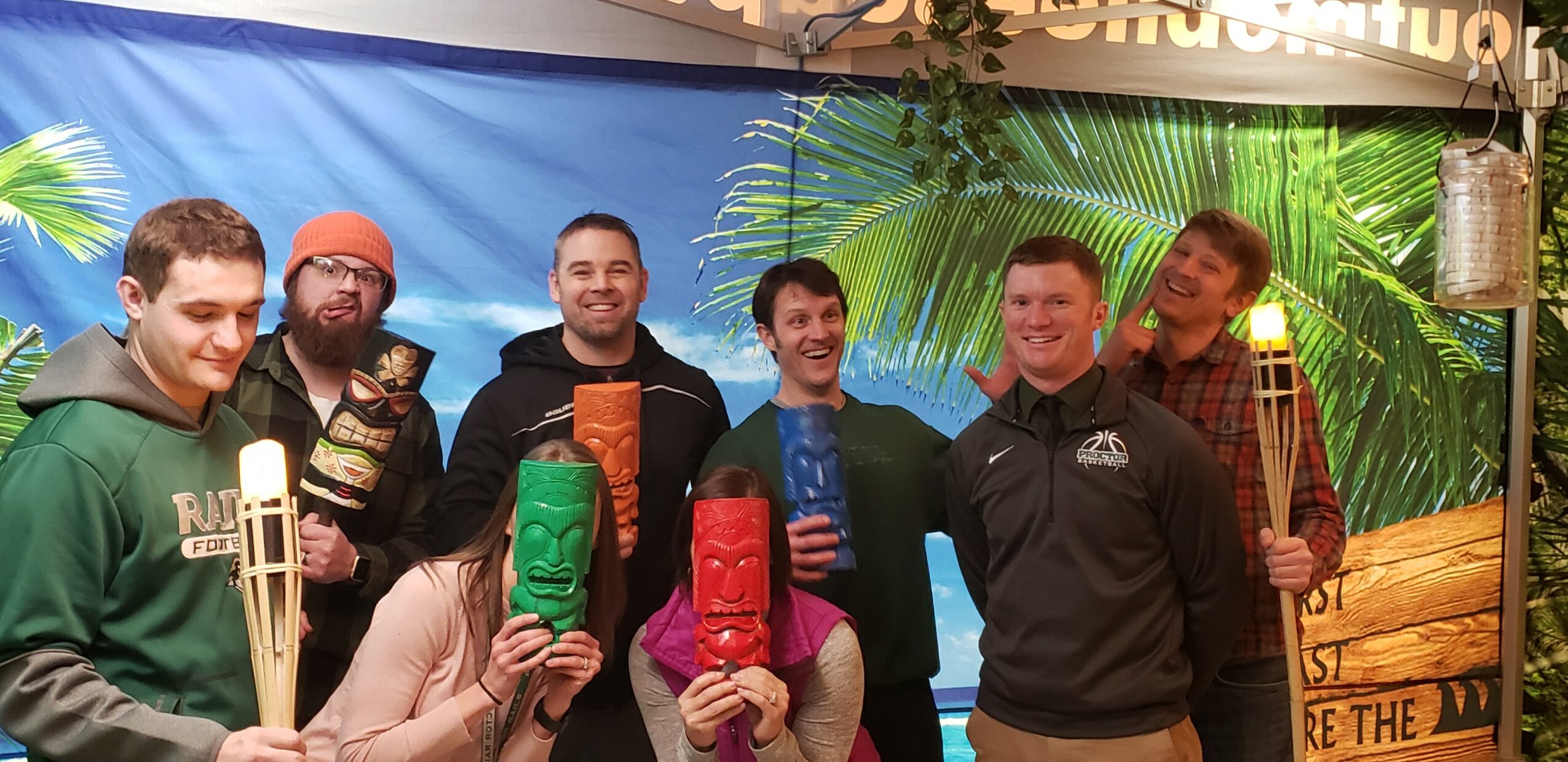 Group of seven people holding tiki masks and torches, standing outdoors with a tropical backdrop, smiling and posing for the photo.