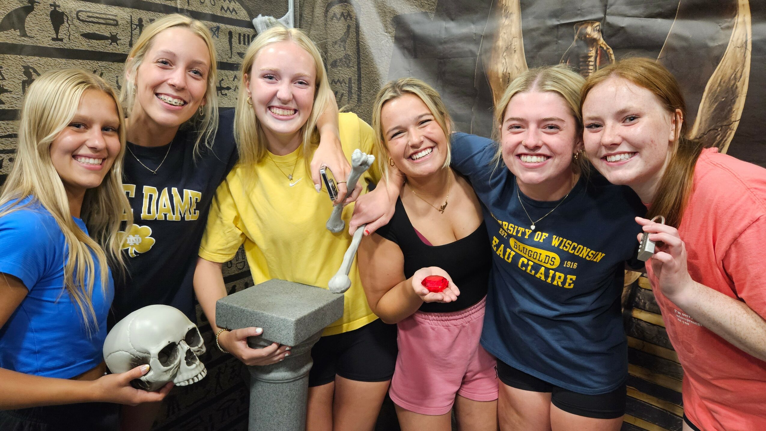 Group of six smiling young women holding skull, hammer, and other objects, standing in front of a dark background with gold accents.