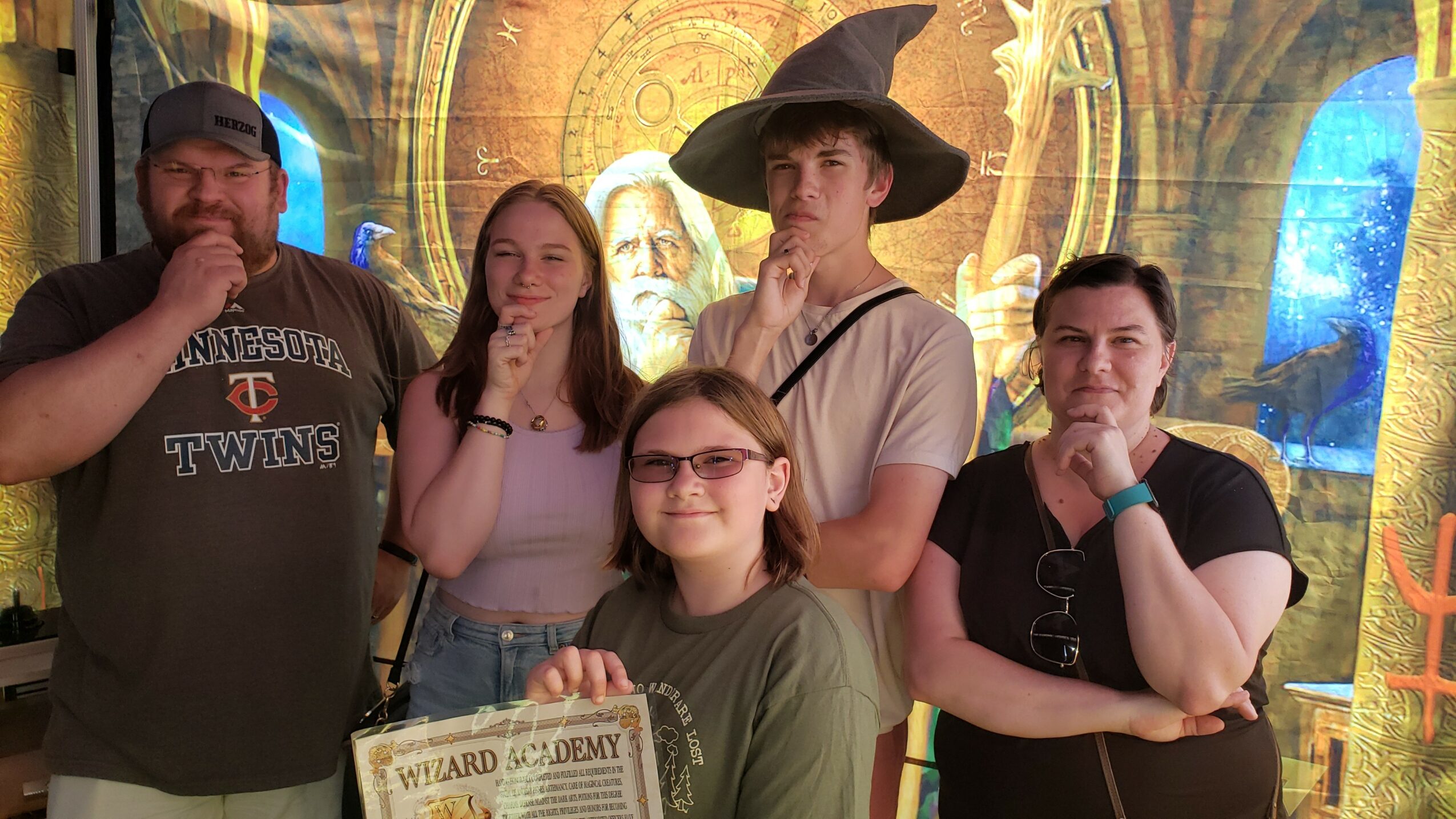 Group of five people posing in front of a colorful background, one dressed as a wizard with a hat, two women, and two men.