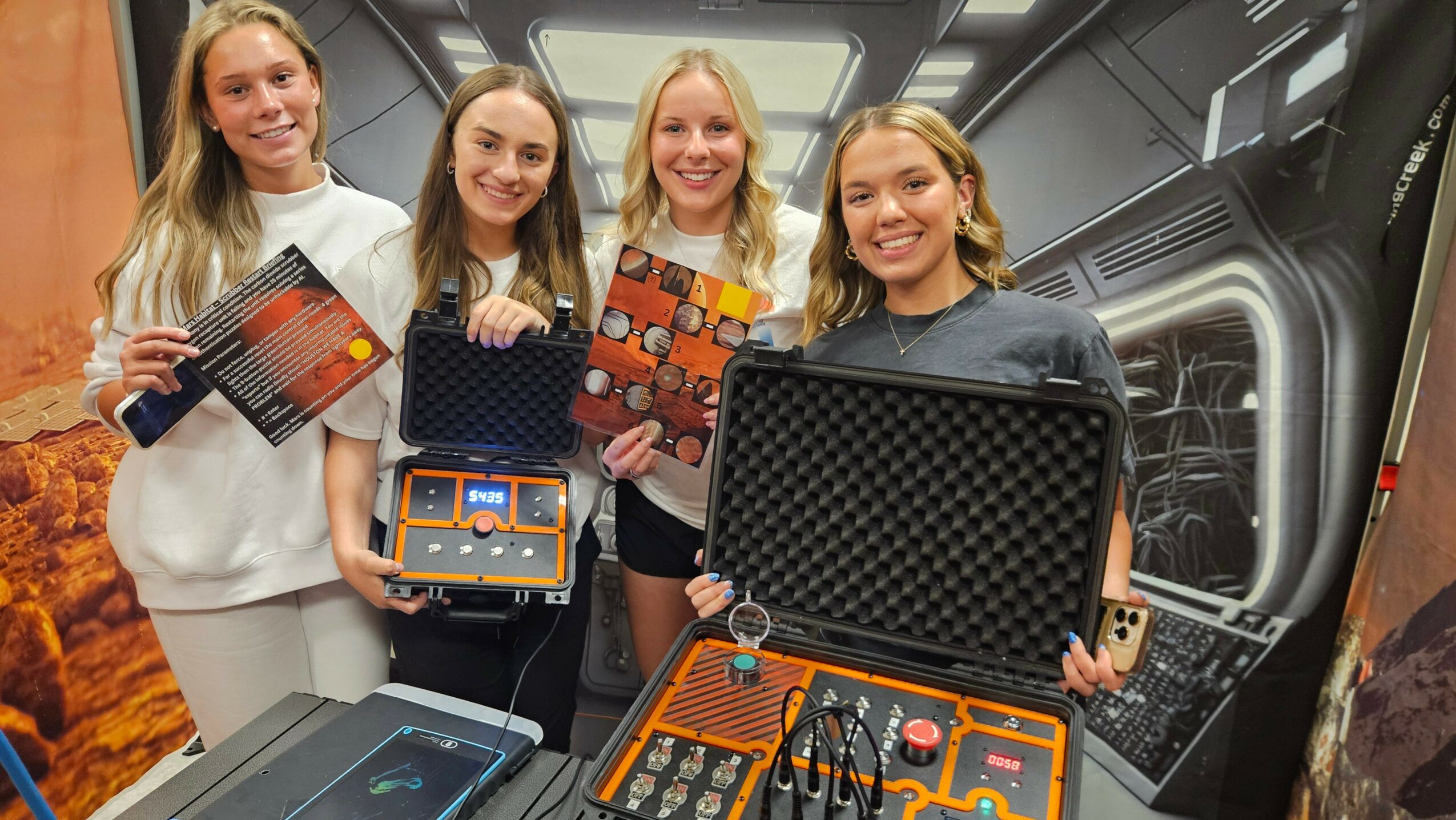 Four smiling women holding electronic equipment cases, standing in front of a sci-fi themed background.
