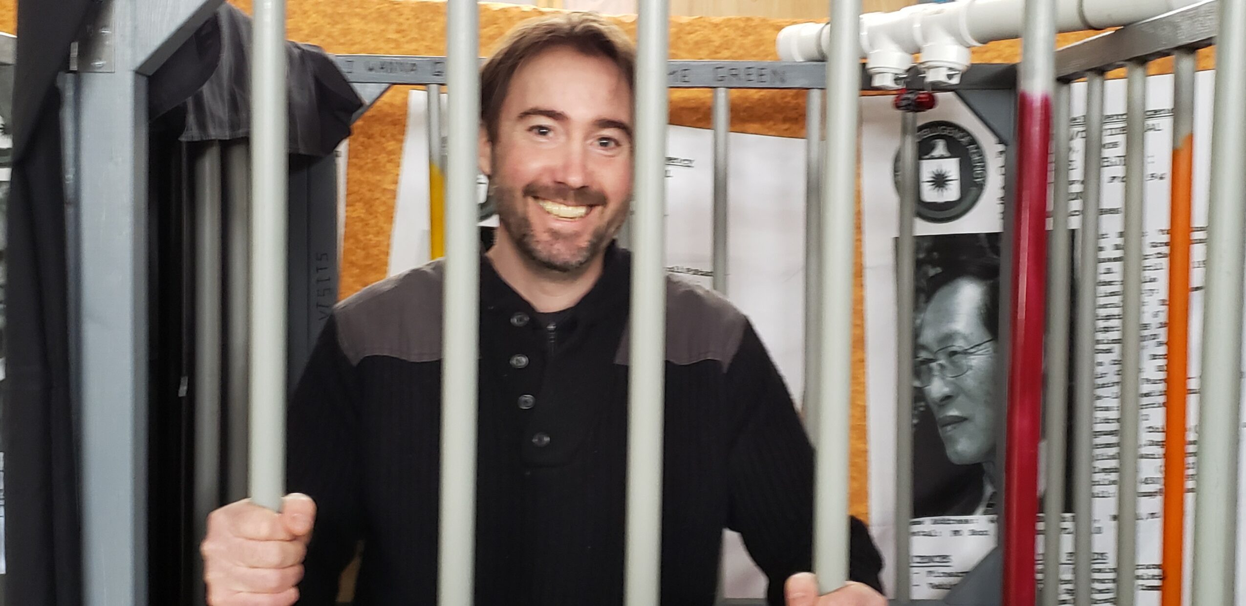 Man smiling behind bars in a cage, with posters and books in the background.