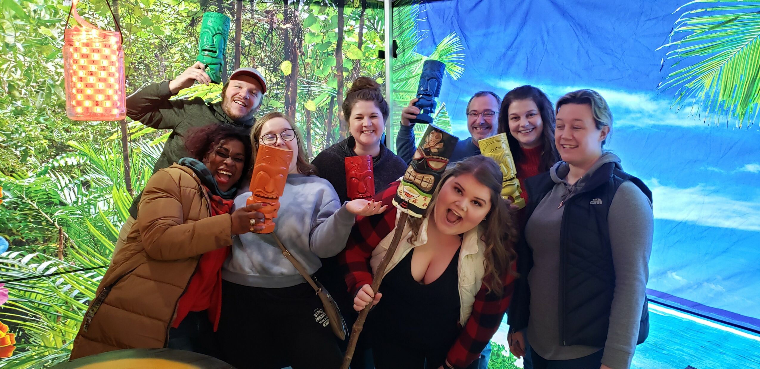 Group of people smiling and holding colorful objects in a jungle-themed setting with blue sky background.