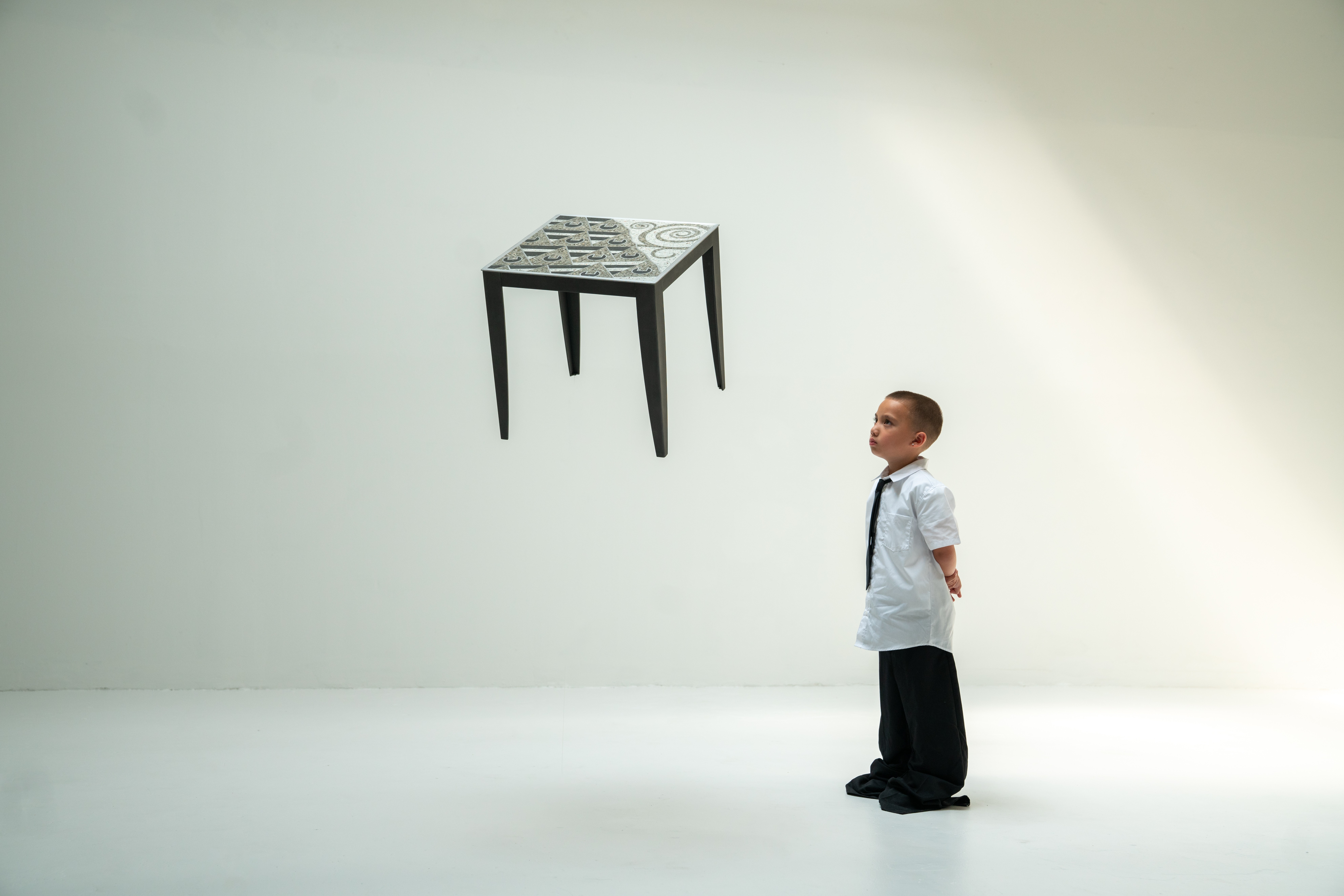 Child in white shirt and dark pants looking at a floating table with a patterned top against a plain background.