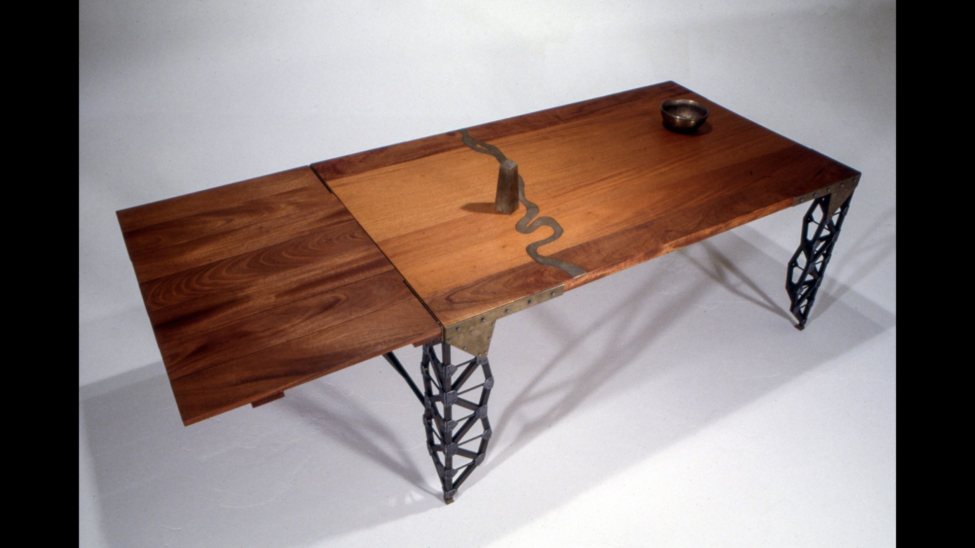 Long wooden table with metal geometric legs, a small bowl, and a decorative object on top, against a plain background.