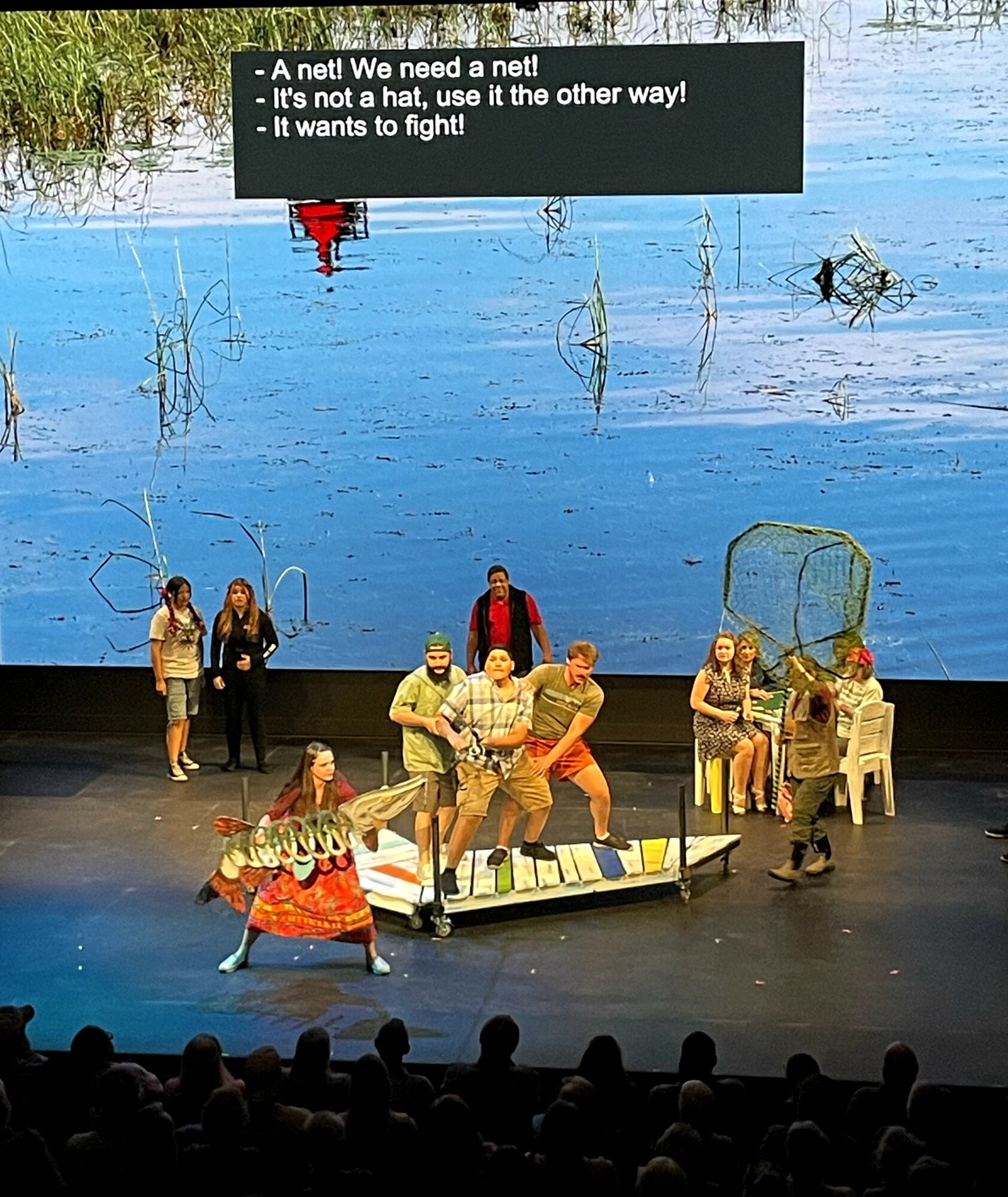 Theater stage with actors performing, some in costumes, and a large screen with text above. Audience visible at bottom.