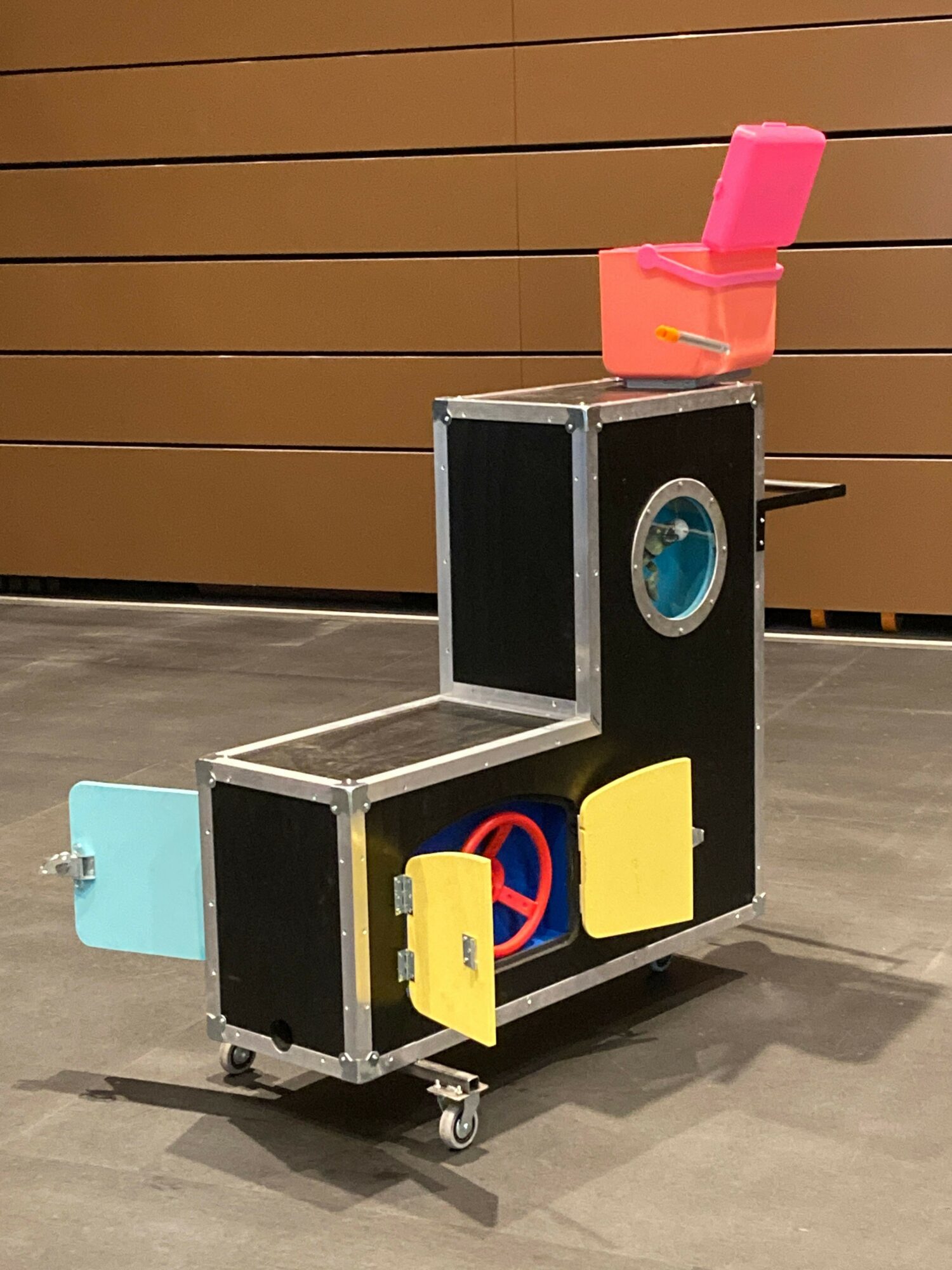 A black and silver box with colorful objects on top, placed on a gray floor against a wooden wall.