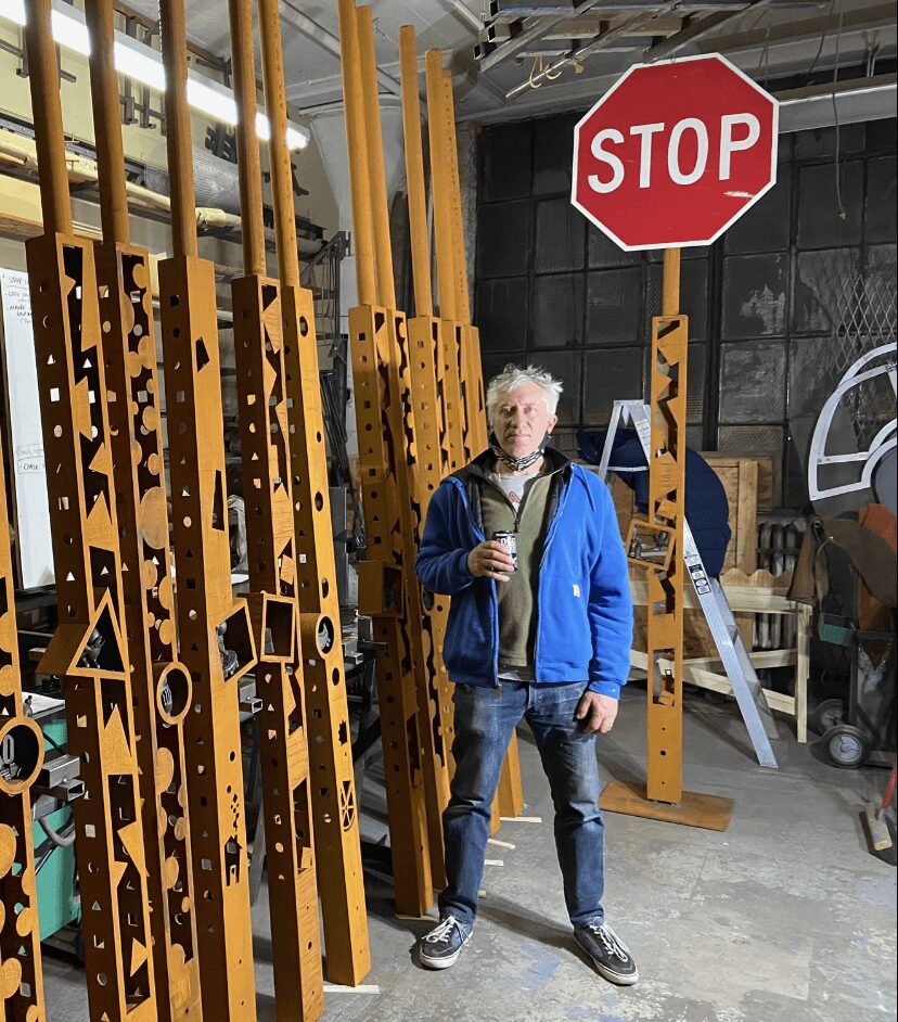 Older man with white hair holding a drink, standing in a warehouse with yellow and black striped objects and a stop sign behind him.