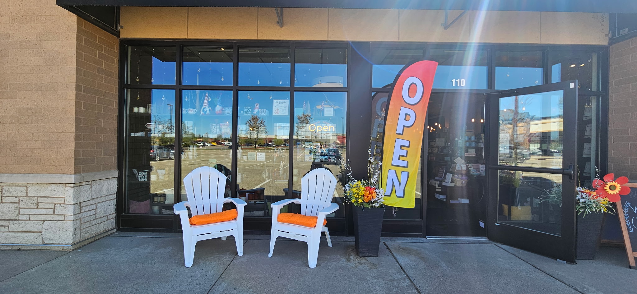 Store entrance with two white chairs, a potted plant, and an open sign, outside glass doors reflecting the sky.