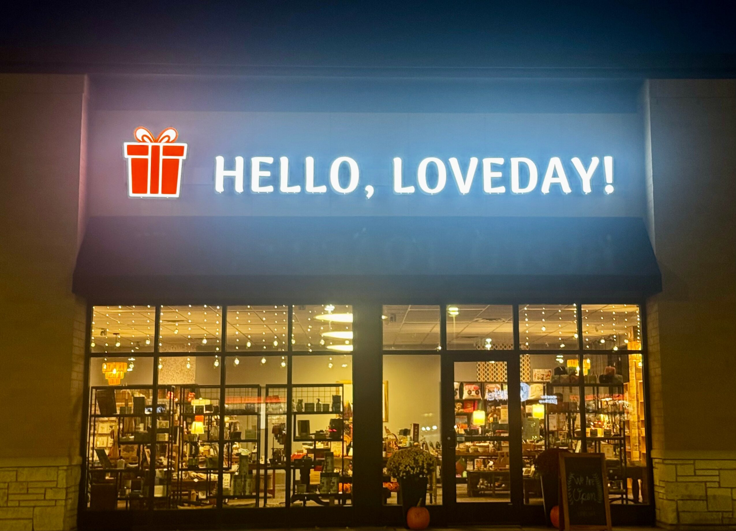 Storefront with illuminated sign reading 'HELLO, LOVEDAY!' and a gift icon, large windows showing interior shelves, night setting.